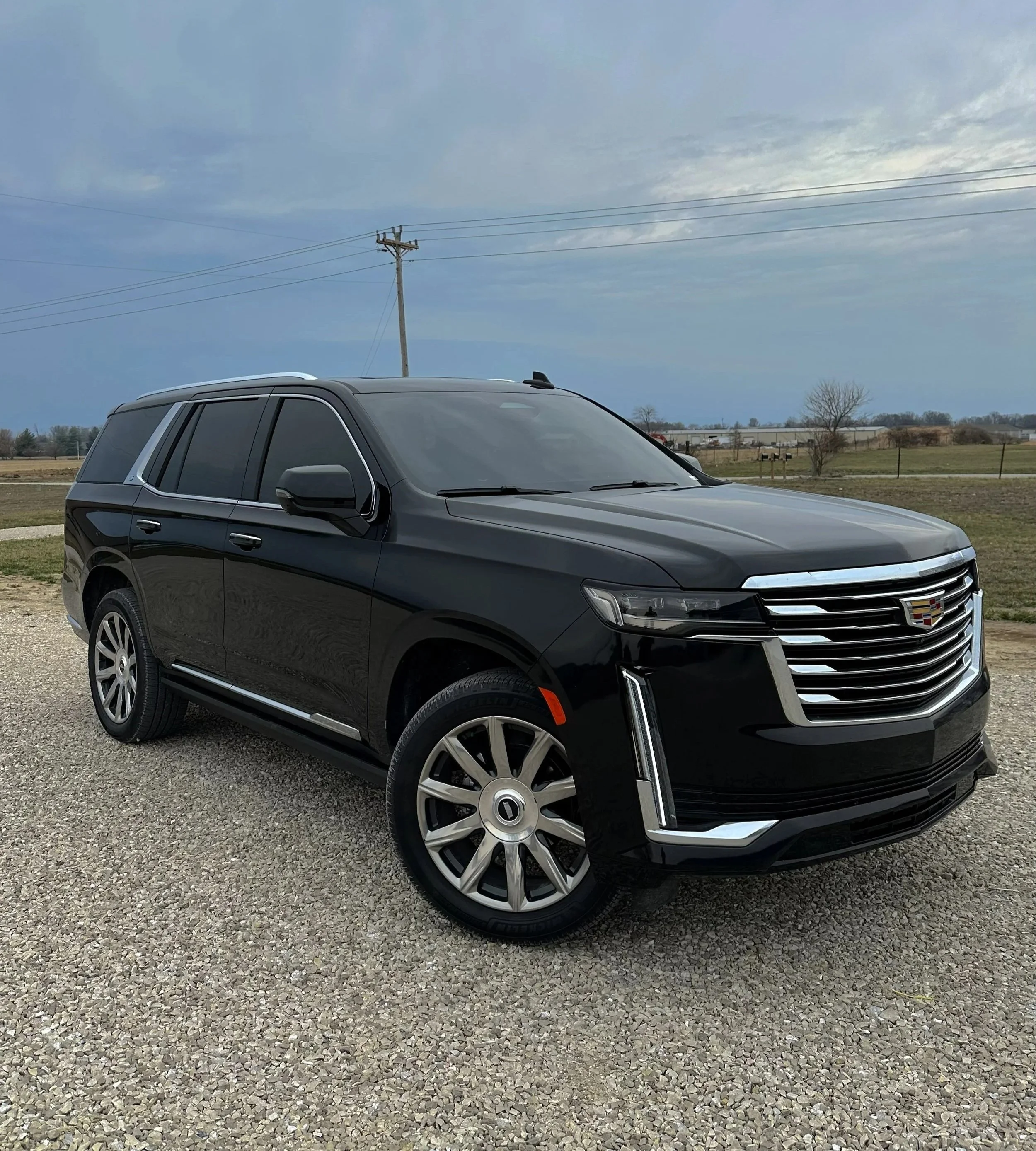 Black Cadillac SUV parked on gravel with open fields and cloudy sky in the background.