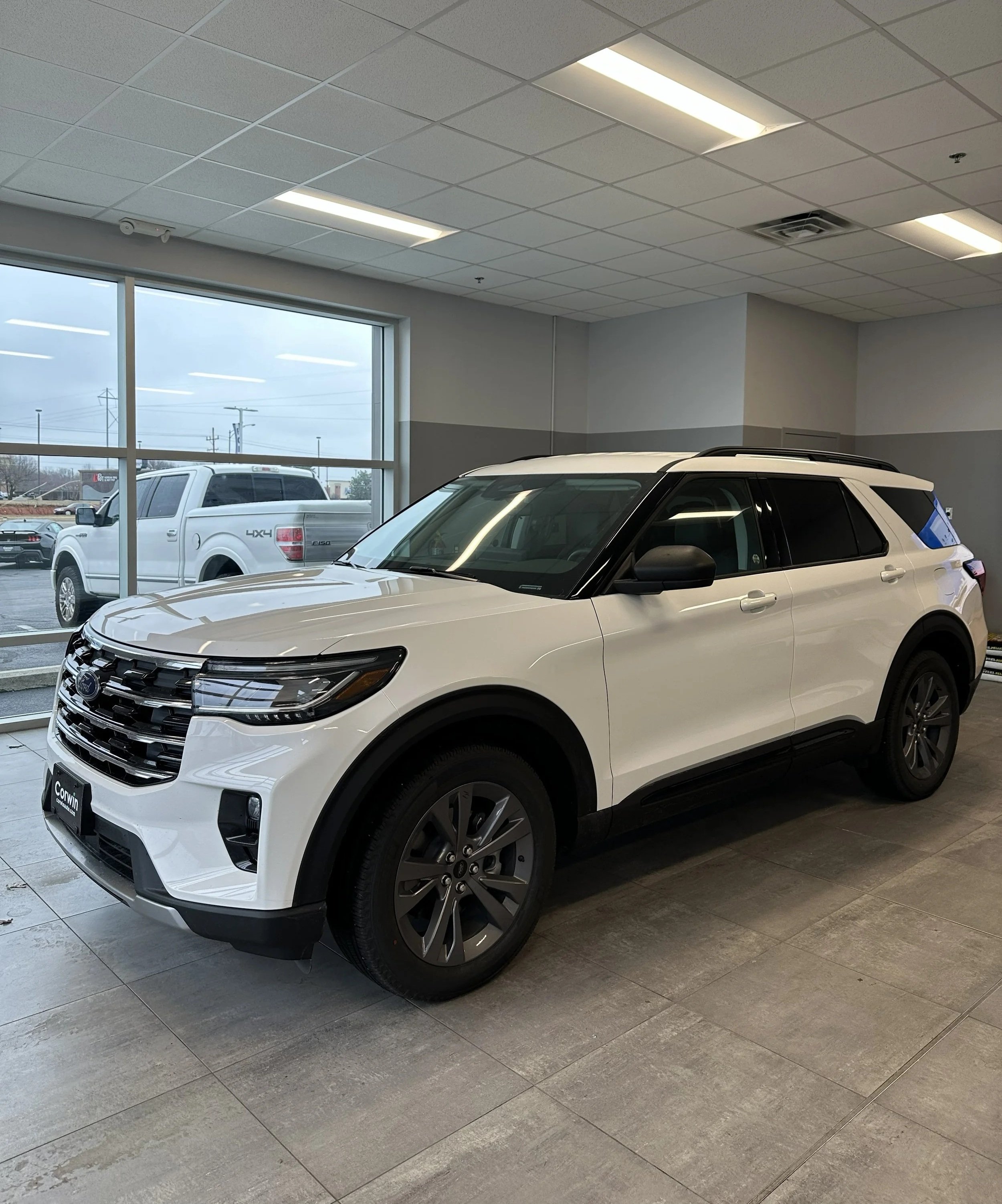 White SUV car displayed inside a showroom with large windows showing parked vehicles outside.