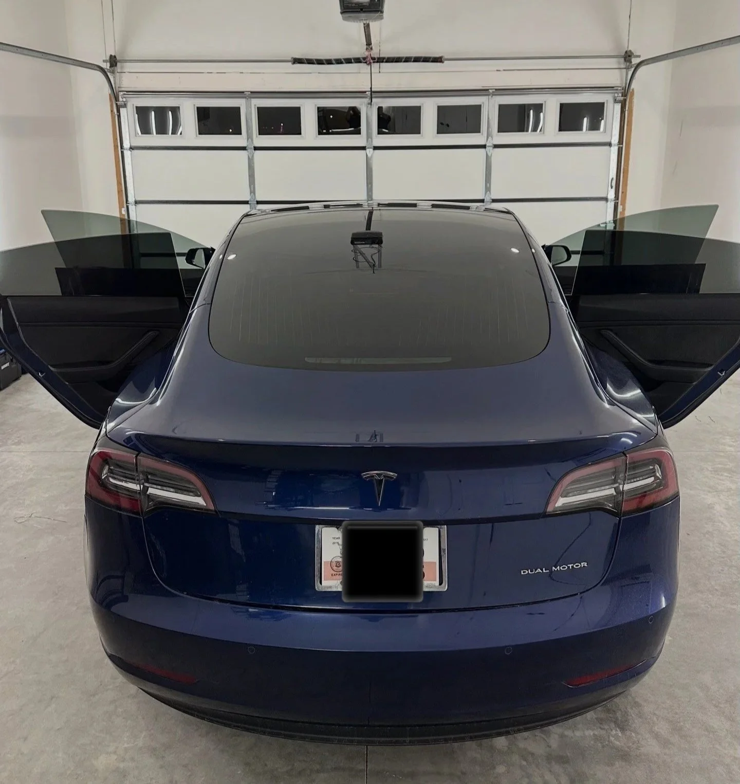 A tinted dark blue Tesla Model 3 with open front doors in a garage, showing the rear windshield and dual motor badge.