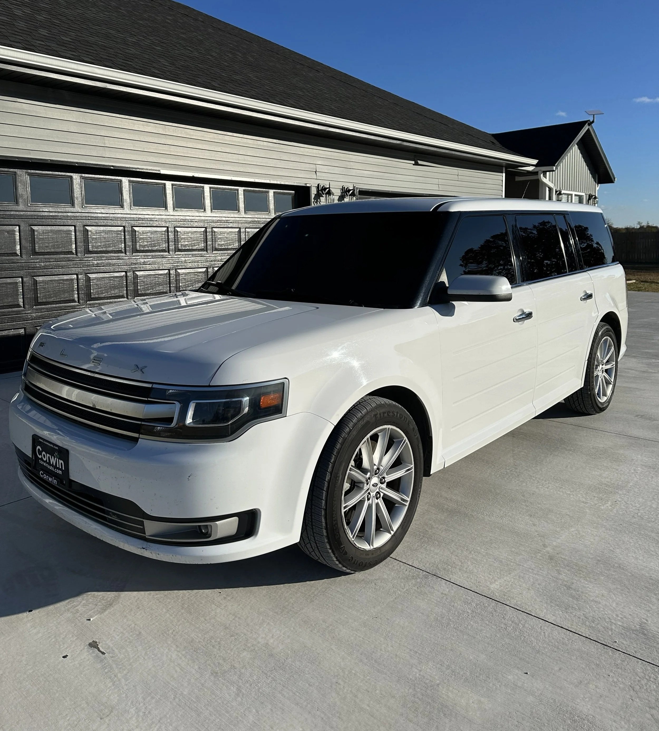 A Tinted Ford Flex on driveway in front of a house with a gray garage door and a black roof, under a blue sky.