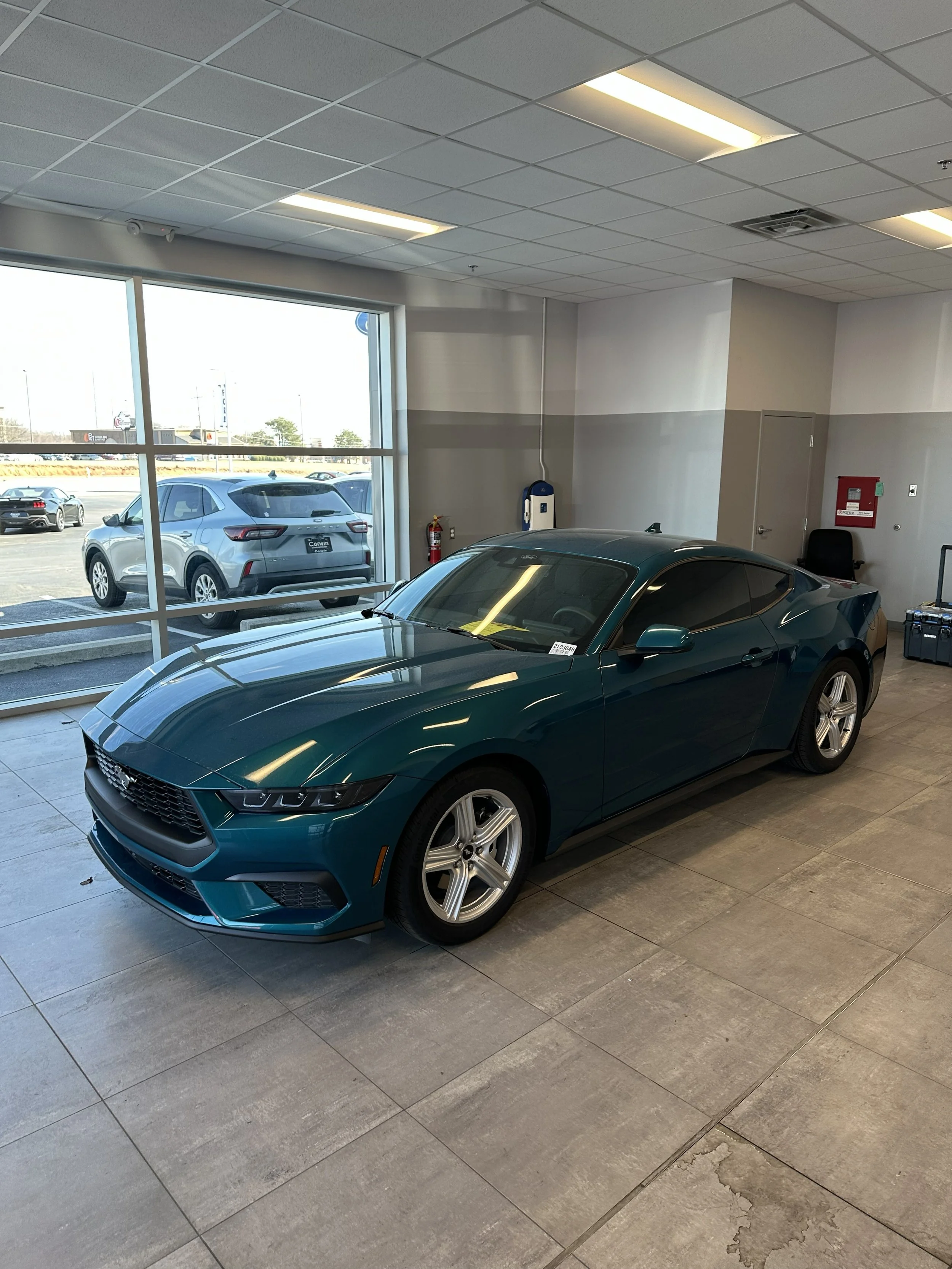 A teal sports car inside a dealership showroom, parked on light-colored tiled flooring, with large windows showing other parked cars outside.