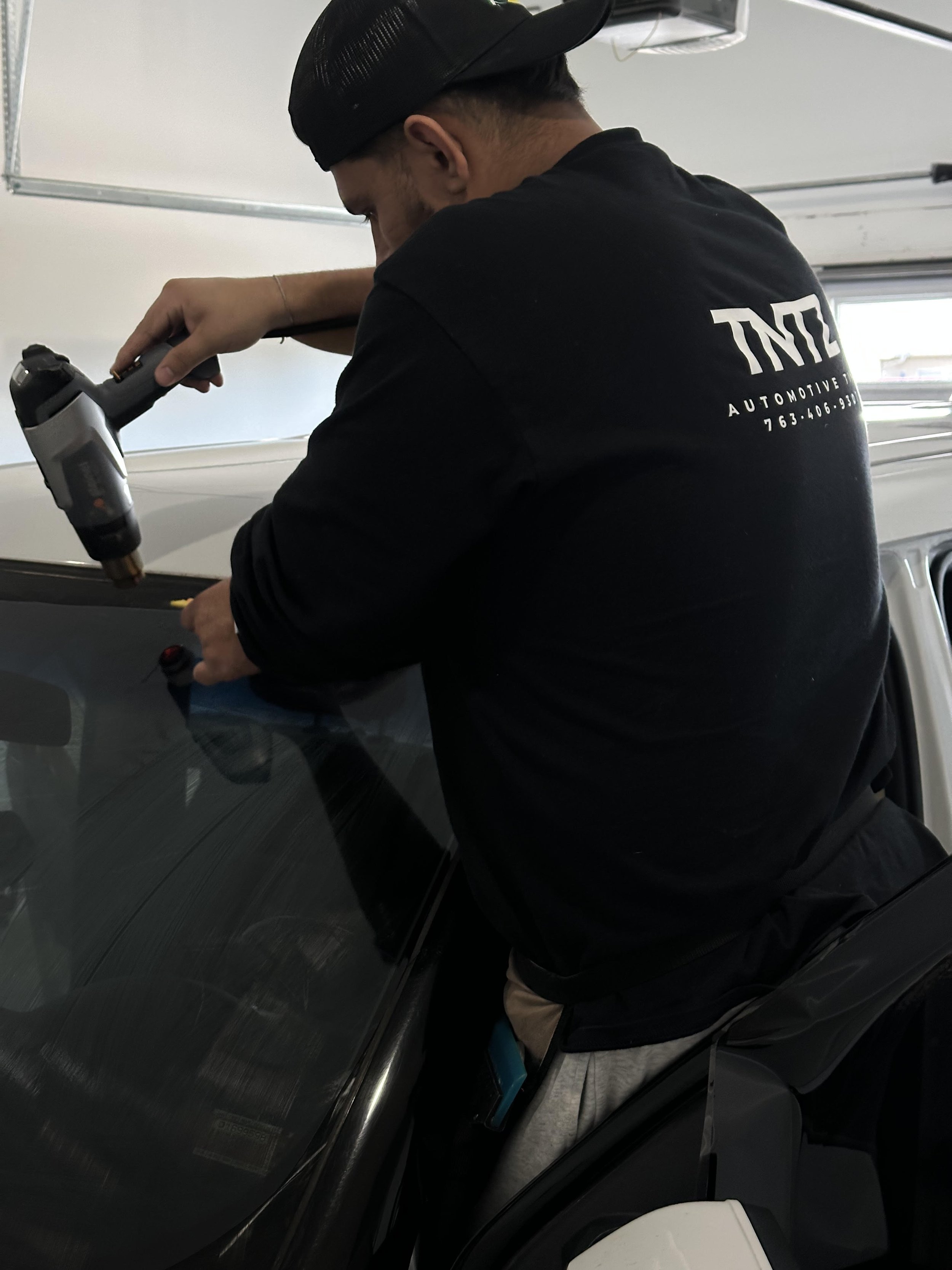 A person using a heat gun on a car's windshield in a garage.