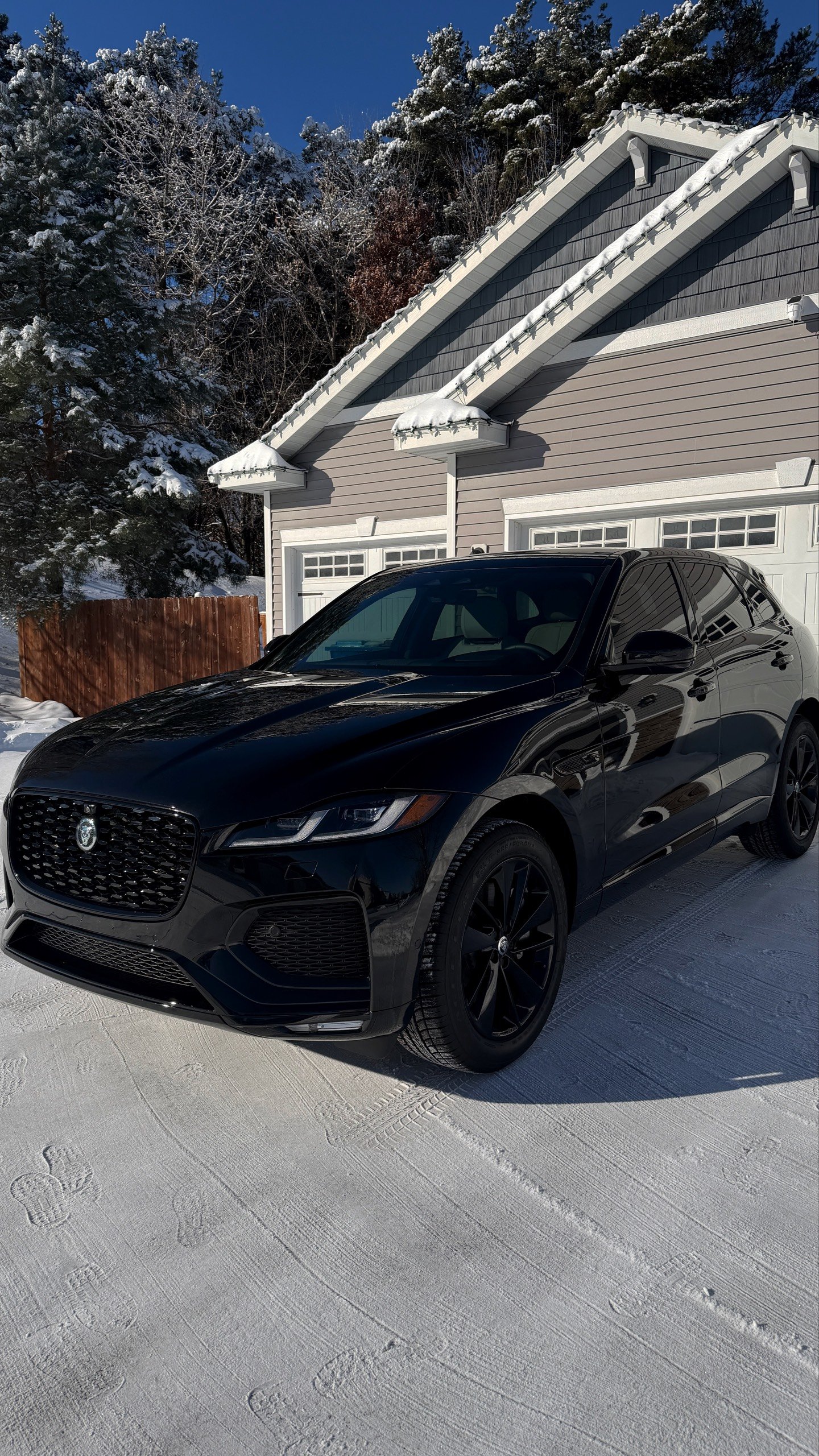 Black Jaguar parked on snowy driveway in front of a house with snow-covered roof and trees in the background.