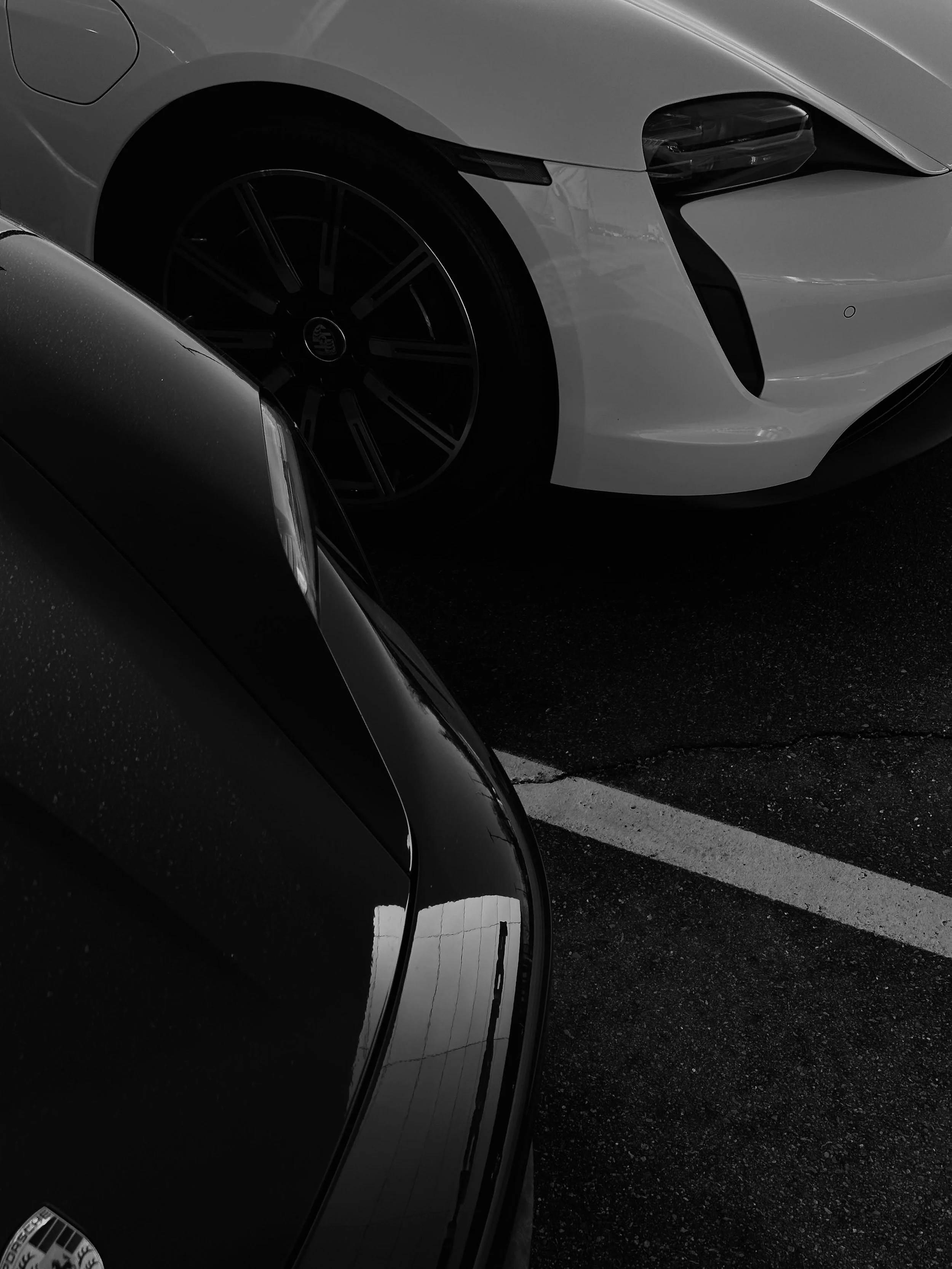 Close-up of front parts of two parked cars, one black with a Porsche logo, and one white with a sleek design, in a parking lot.