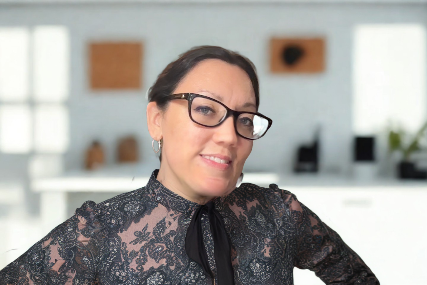 A woman with short dark hair, glasses, and hoop earrings smiling at the camera in an office setting with blurred decor and desks in the background.