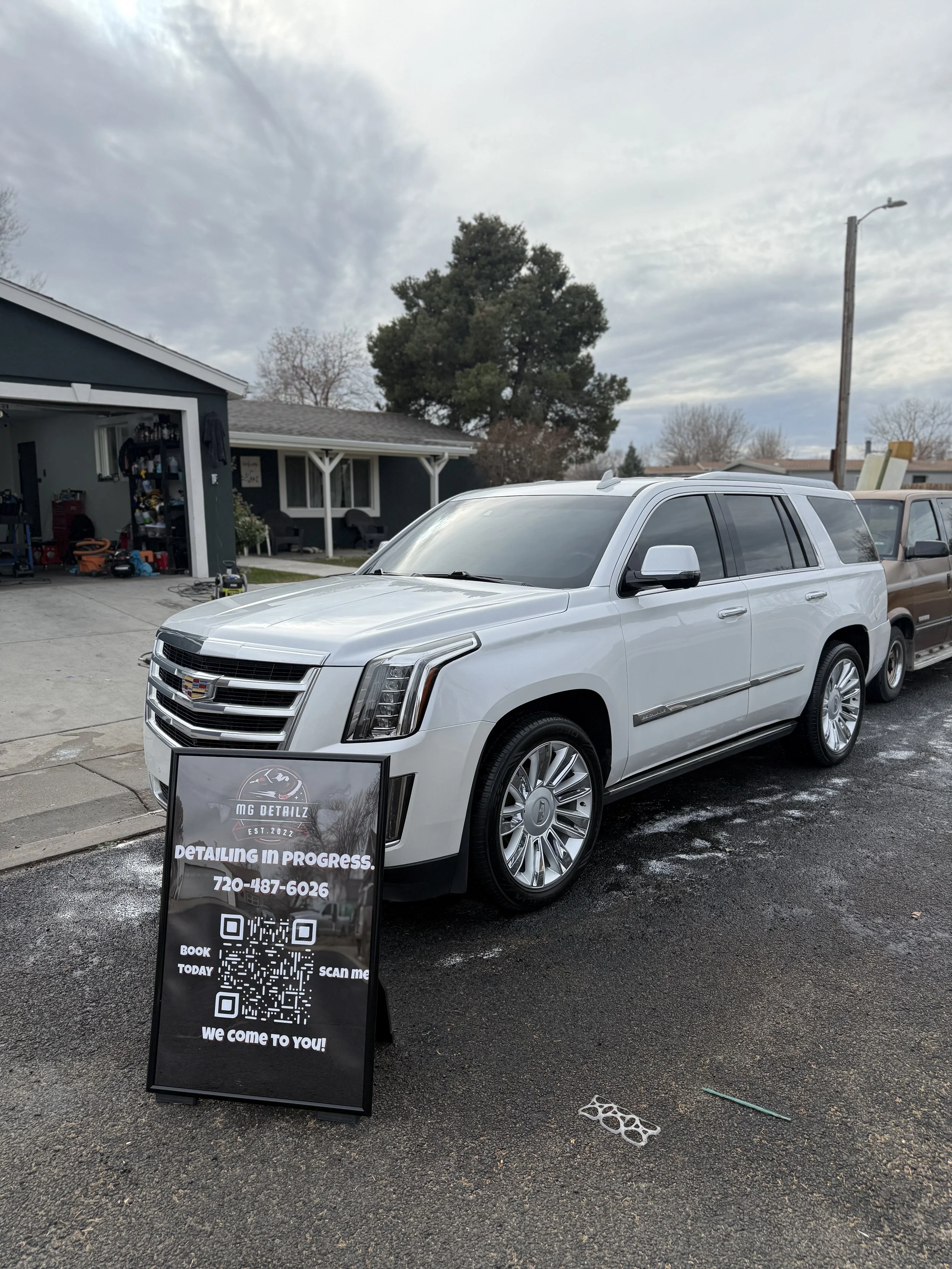 White Cadillac SUV parked on a street next to a sign for a detailing service that reads 'MG Detailing, Detailing in Progress, 720-487-6026, Book Today, Scan Me, We Come to You!' with QR codes. Residential houses are visible in the background under a 