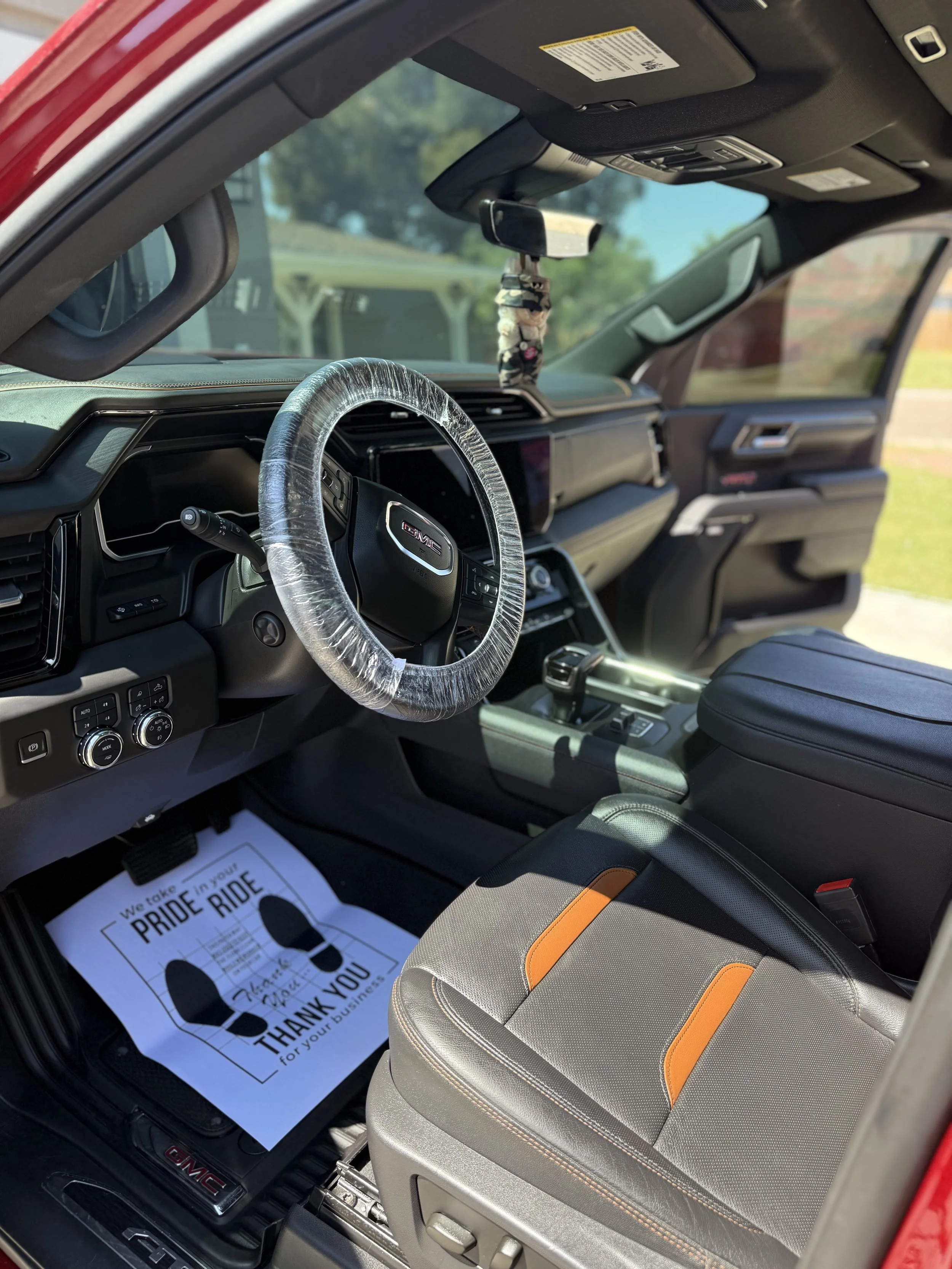 Interior of a GMC vehicle with a plastic-wrapped steering wheel, a paper floor mat thanking for business, and a black dashboard with a hanging decoration from the rearview mirror.
