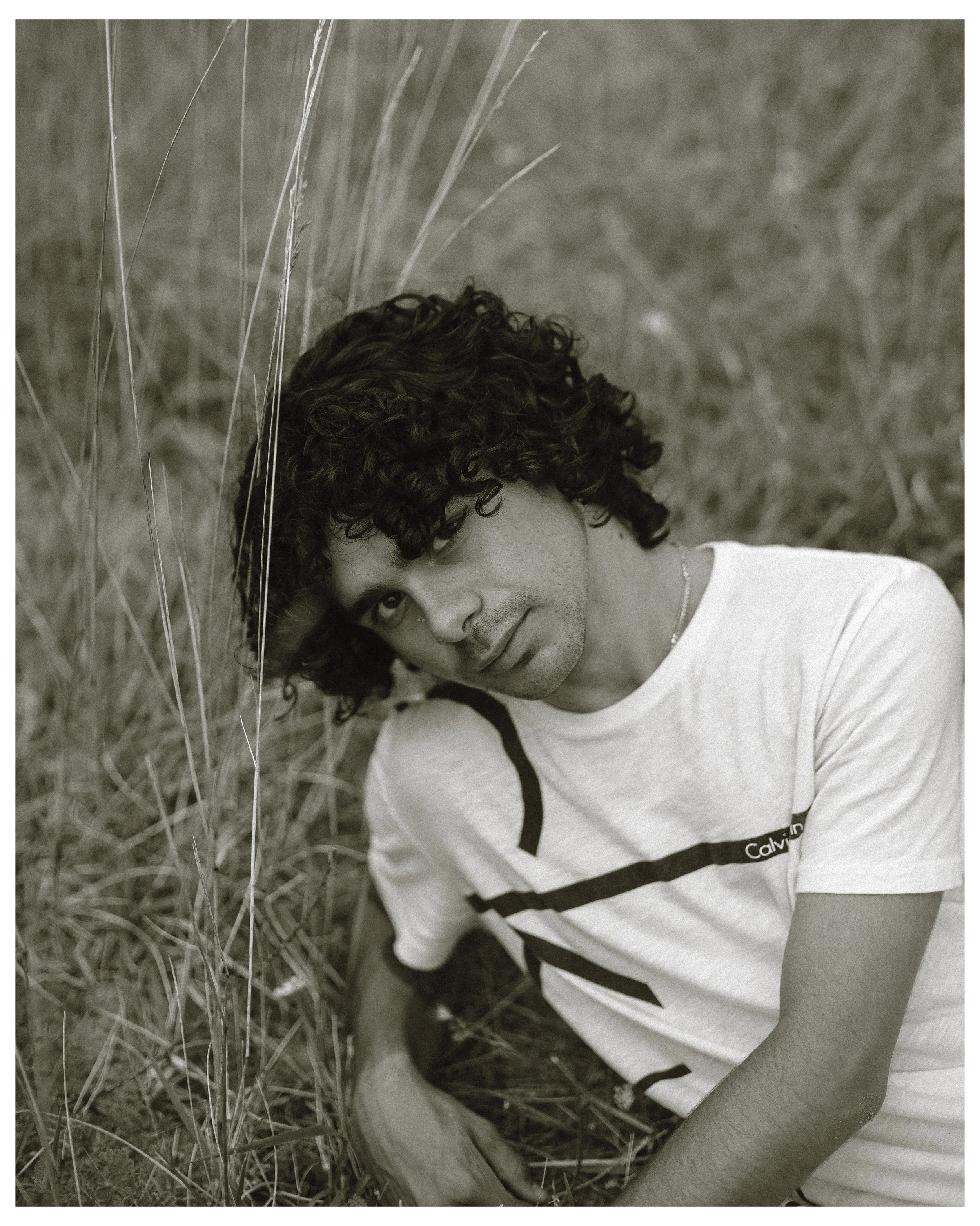 A young man with curly hair lying on the ground in a field of tall grass, looking at the camera.