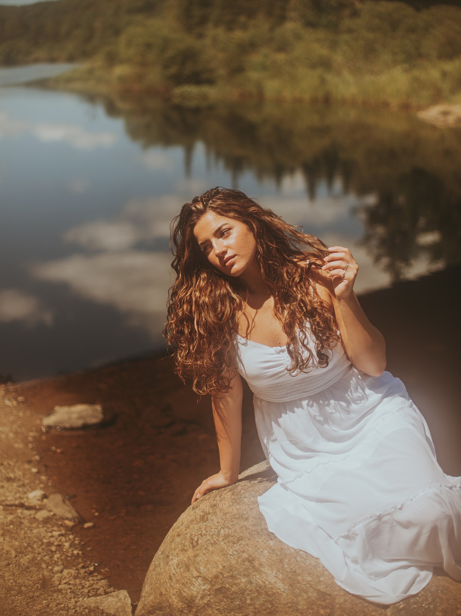 A woman with long curly hair in a white dress sits on a large rock near a calm body of water, with trees and sky reflected in the water.