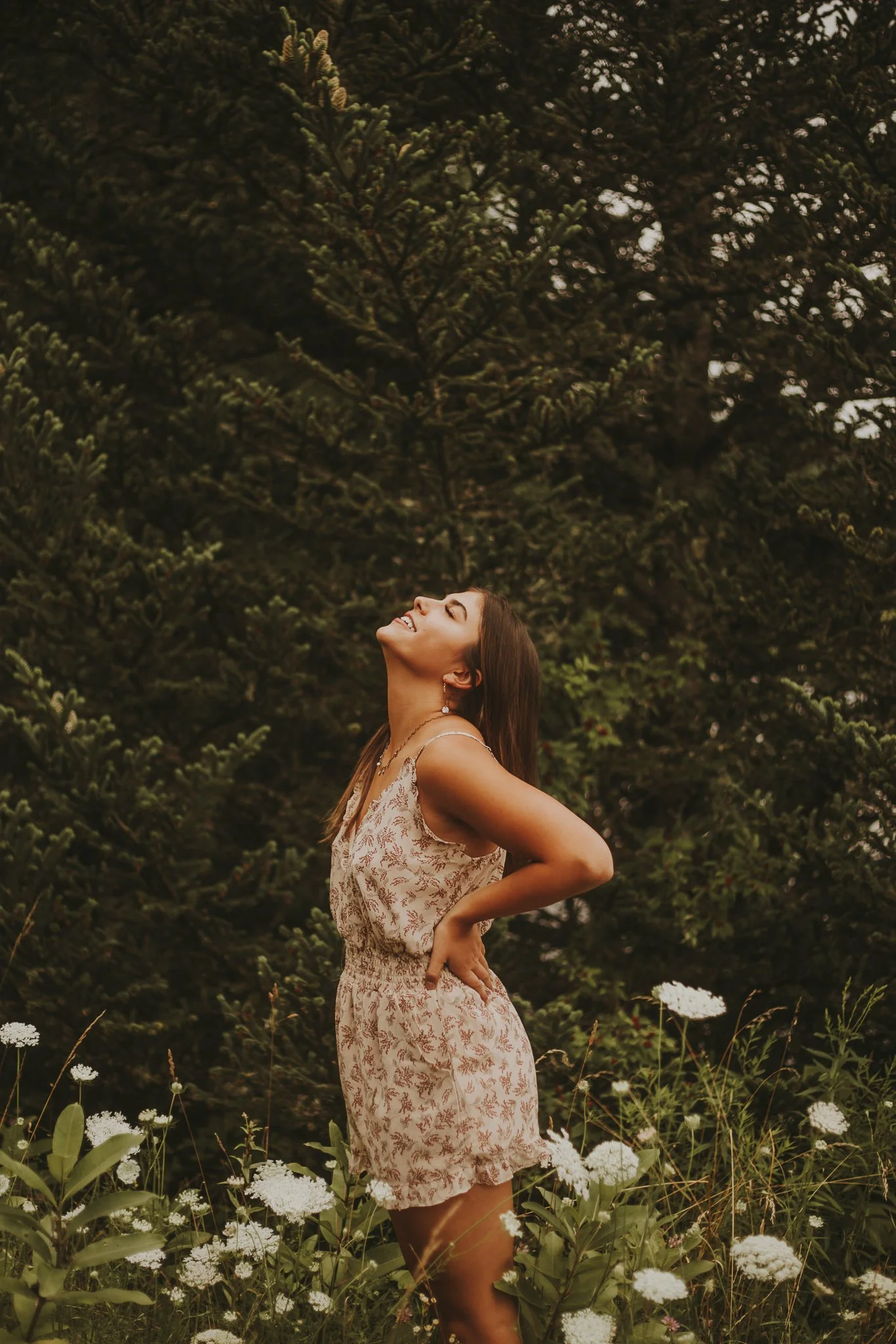 A woman standing outdoors near white flowering plants, with a large green pine tree in the background. She is smiling and tilting her head back with her eyes closed, wearing a floral dress.