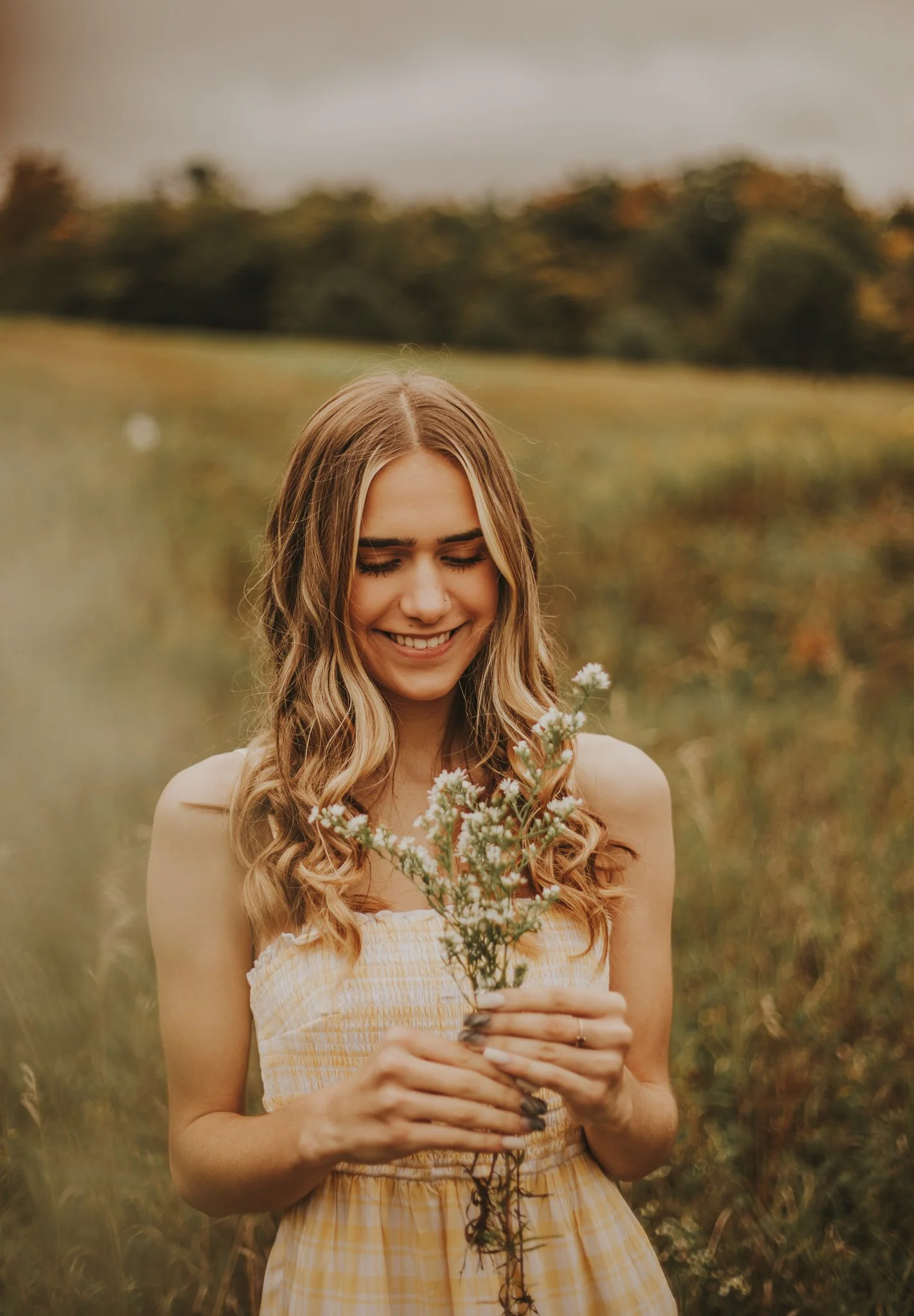 A young woman with long wavy hair smiling while holding a small bouquet of white flowers in a grassy field.