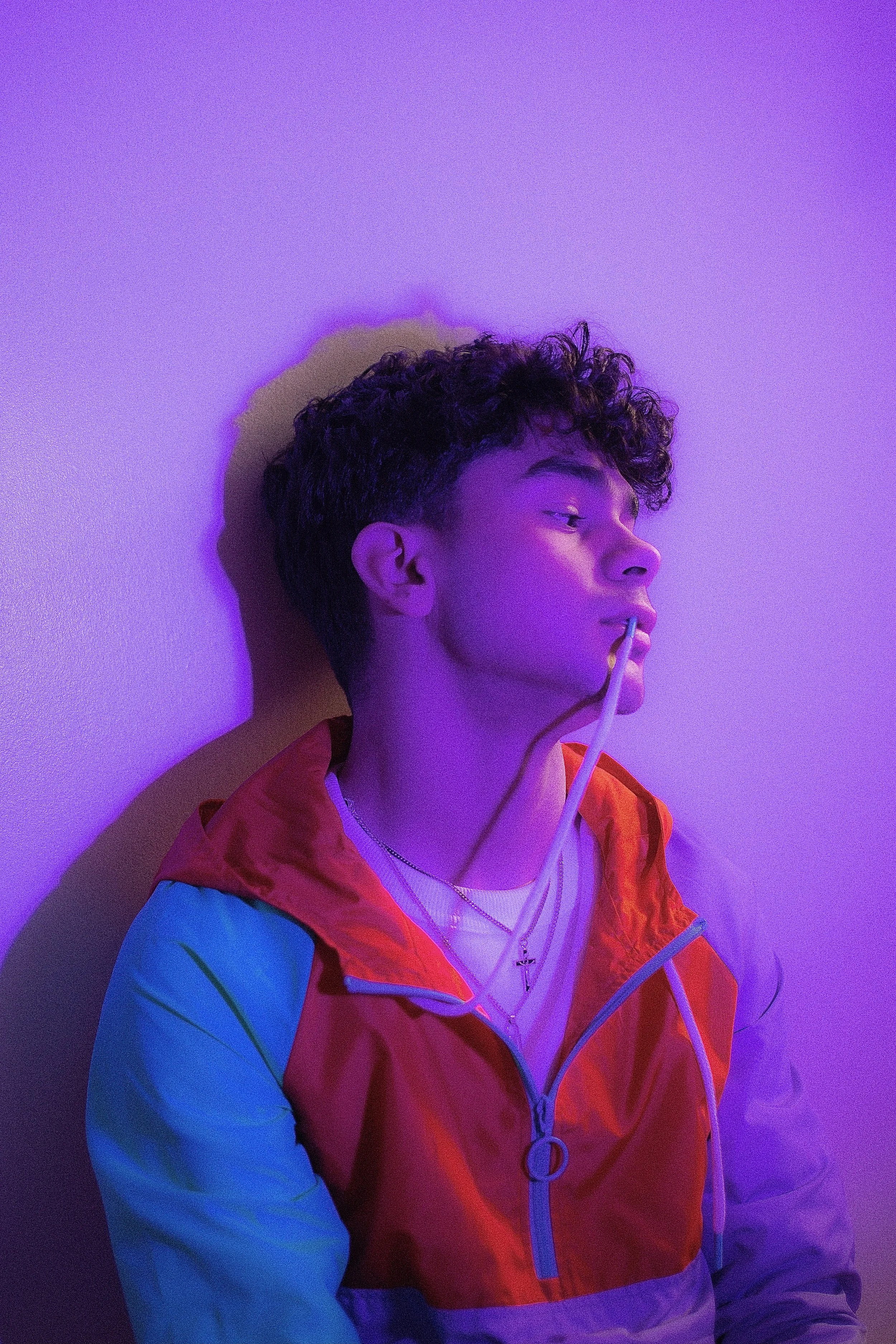 A young man with curly hair leaning against a purple wall, wearing a colorful jacket and holding earbuds in his mouth, illuminated by purple lighting.