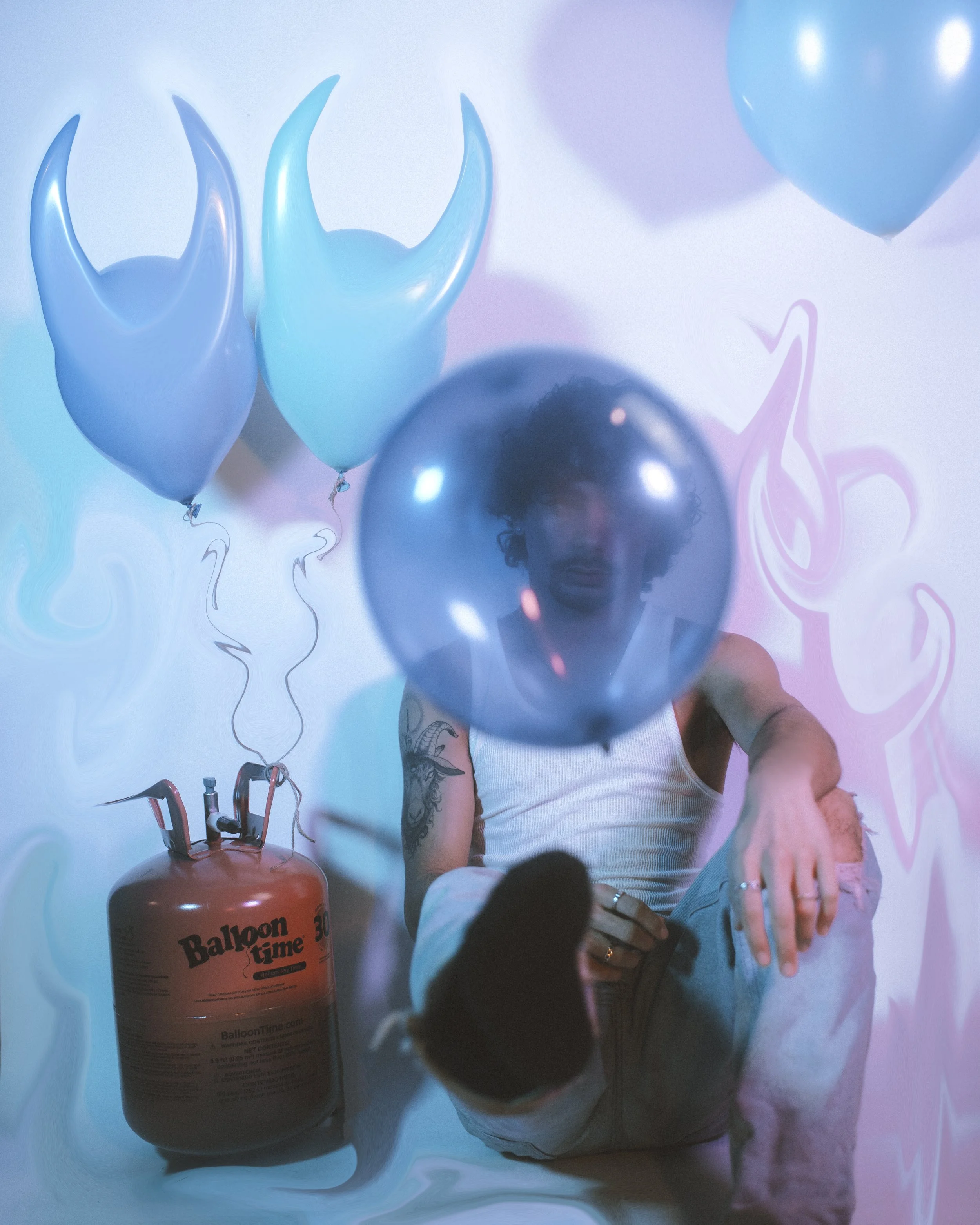 Person sitting on the floor with a gas cylinder, surrounded by blue and pink balloons, with their face reflected in a blue balloon, in a colorful party setting.