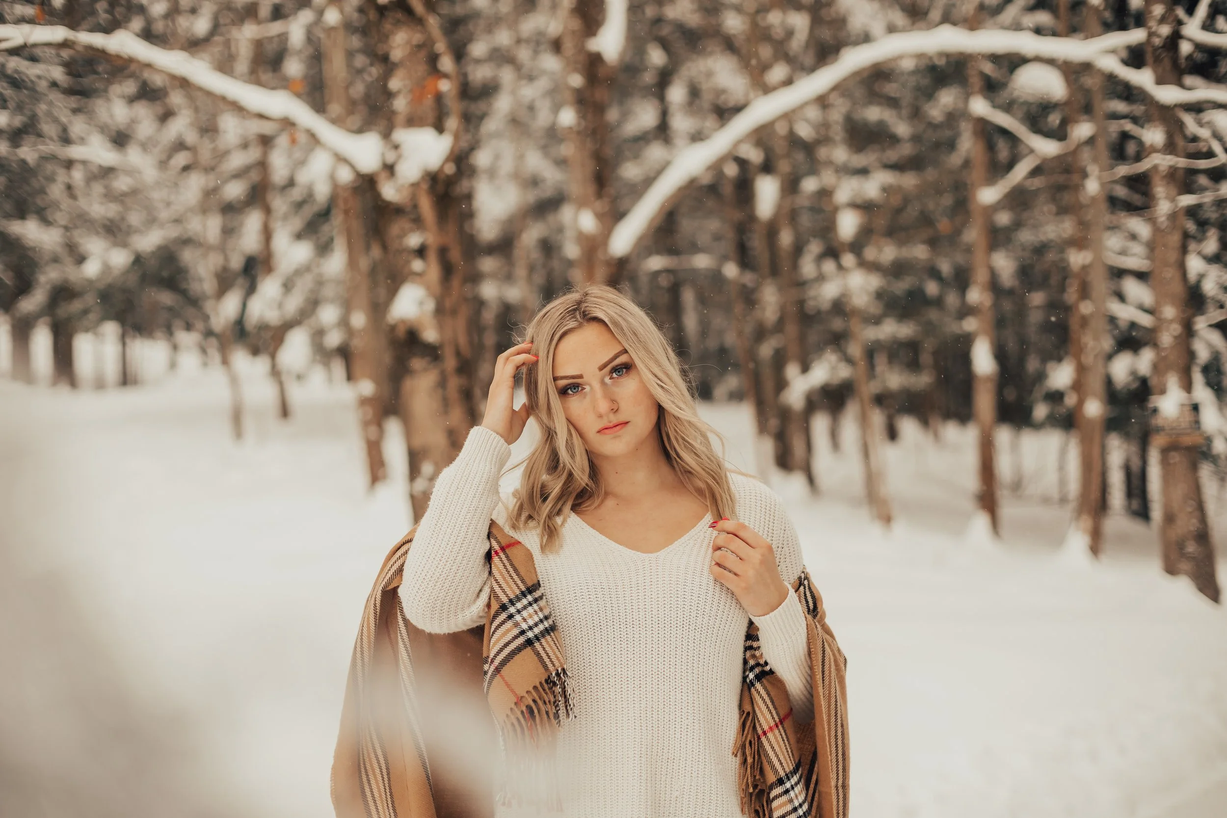 A young woman with blonde hair, wearing a white sweater and a tan plaid coat, stands in a snowy forest with tall trees covered in snow, looking directly at the camera with her hand touching her hair.