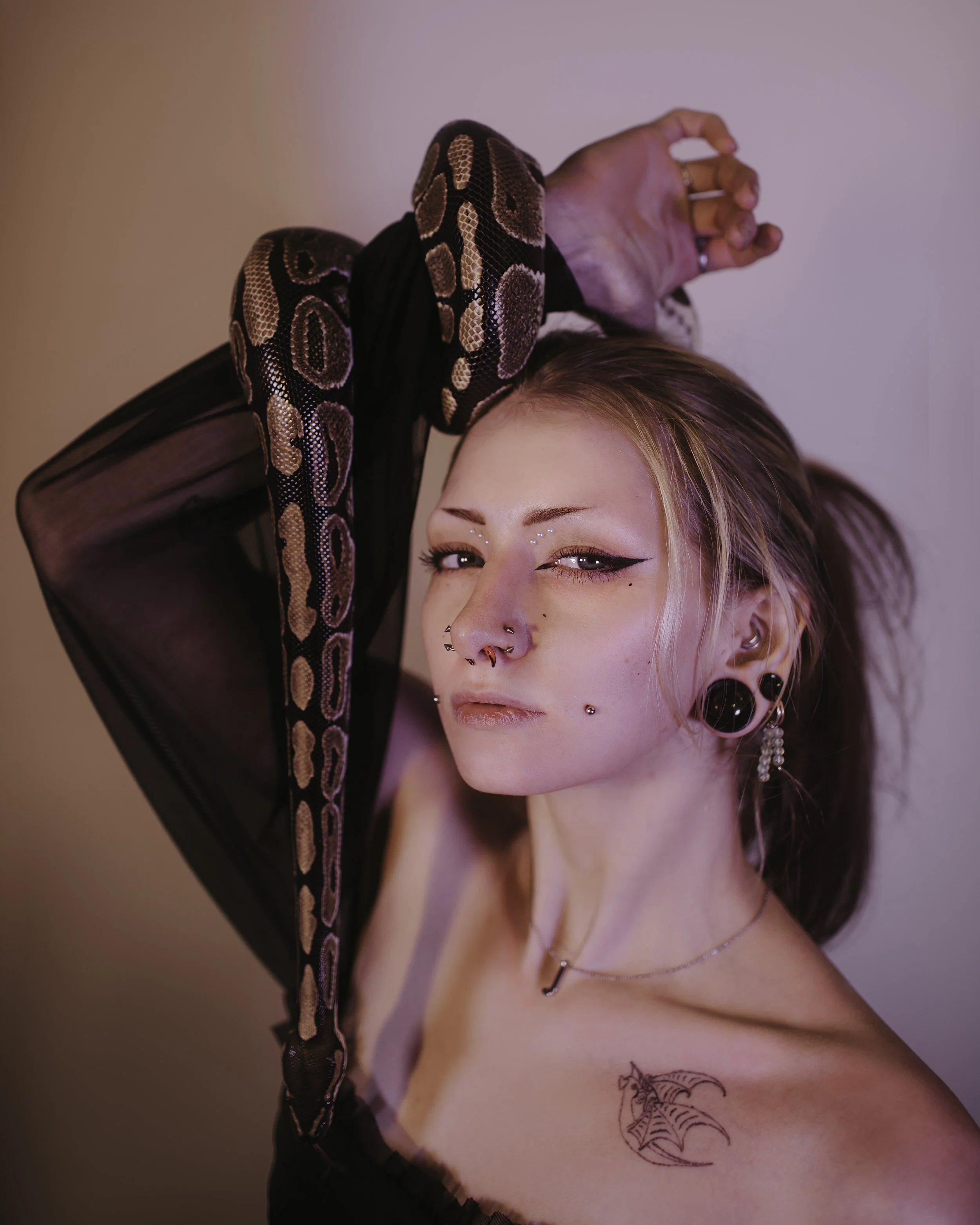 A young woman with earrings, piercings, and a tattoo, holding a snake over her shoulder, with a confident expression.