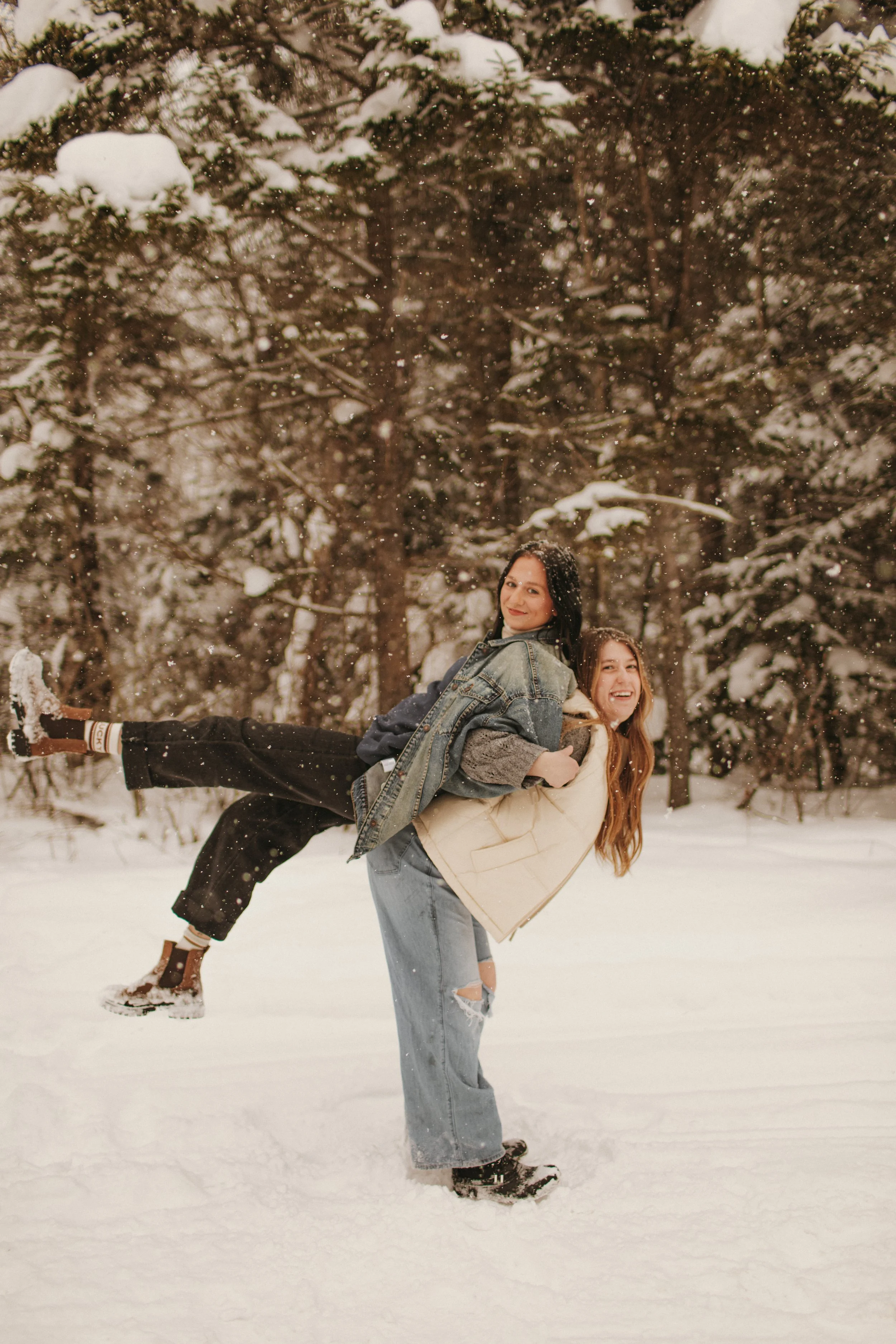 Two young women are in a snowy forest, with one person giving the other a piggyback ride. Both are smiling and dressed in winter clothing, surrounded by snow-covered trees.