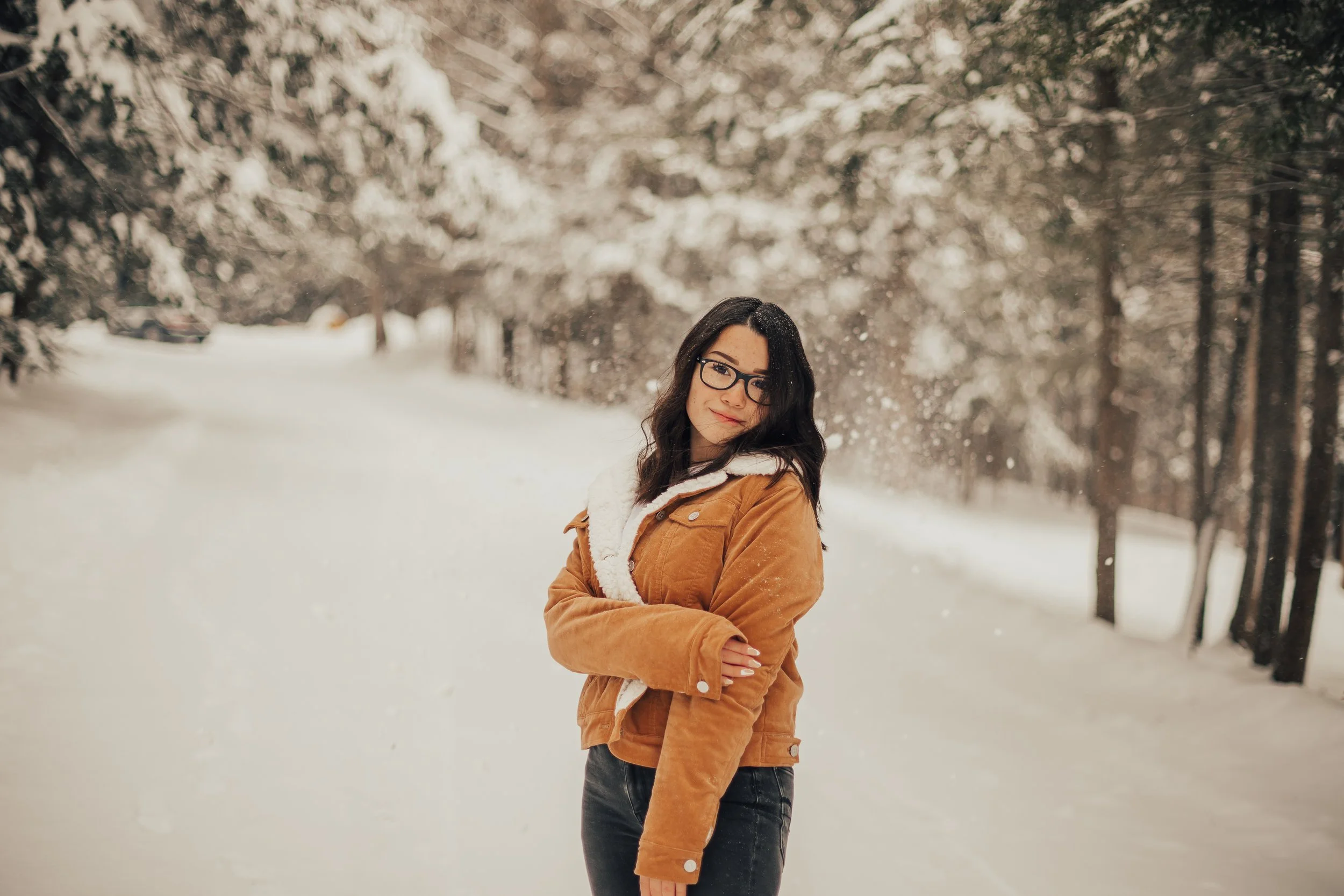 A woman with glasses and dark hair, wearing a tan jacket, standing outdoors in a snow-covered forest.