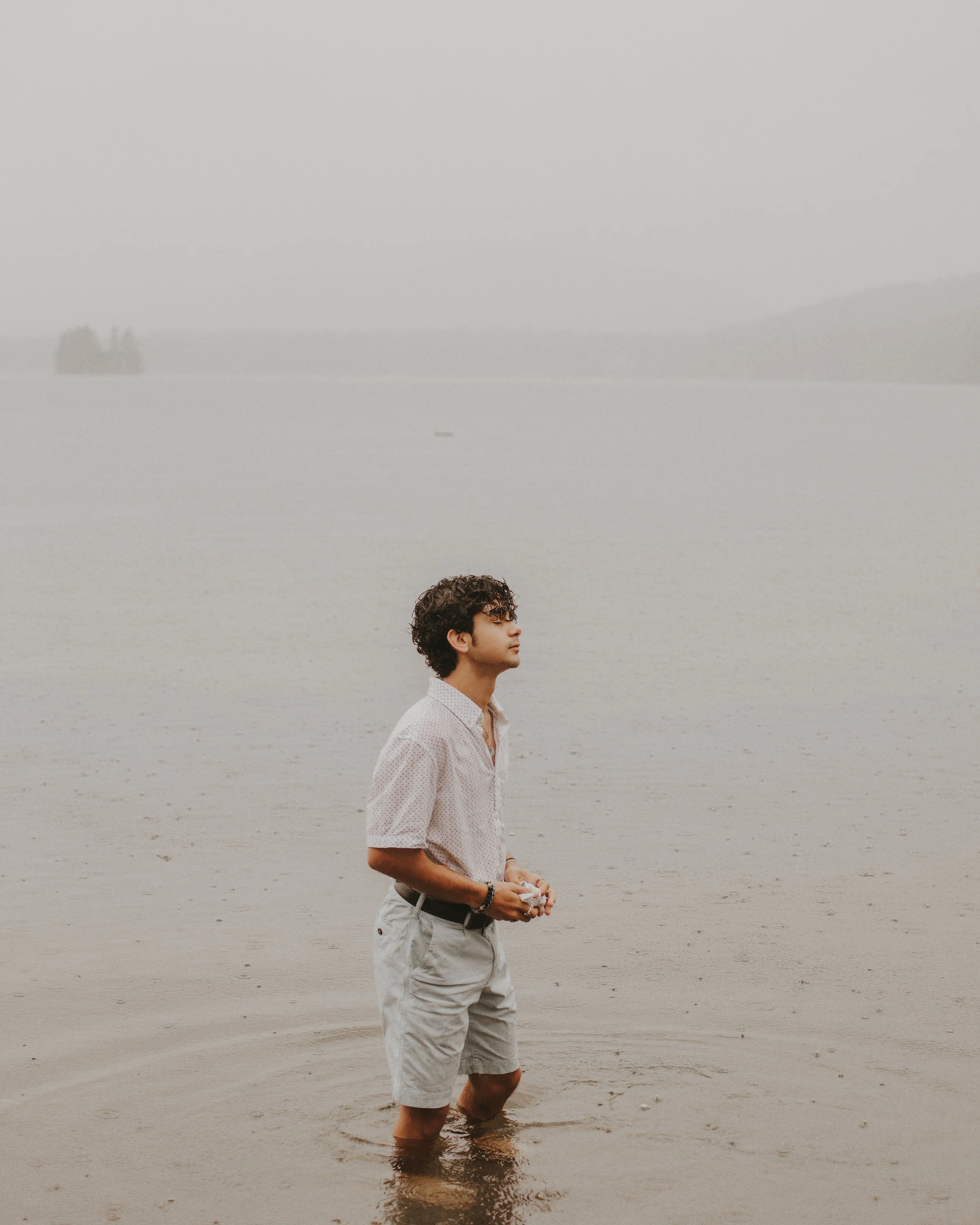 A young man standing in shallow water at the beach, appearing to be deep in thought, with a foggy background and distant trees on the horizon.