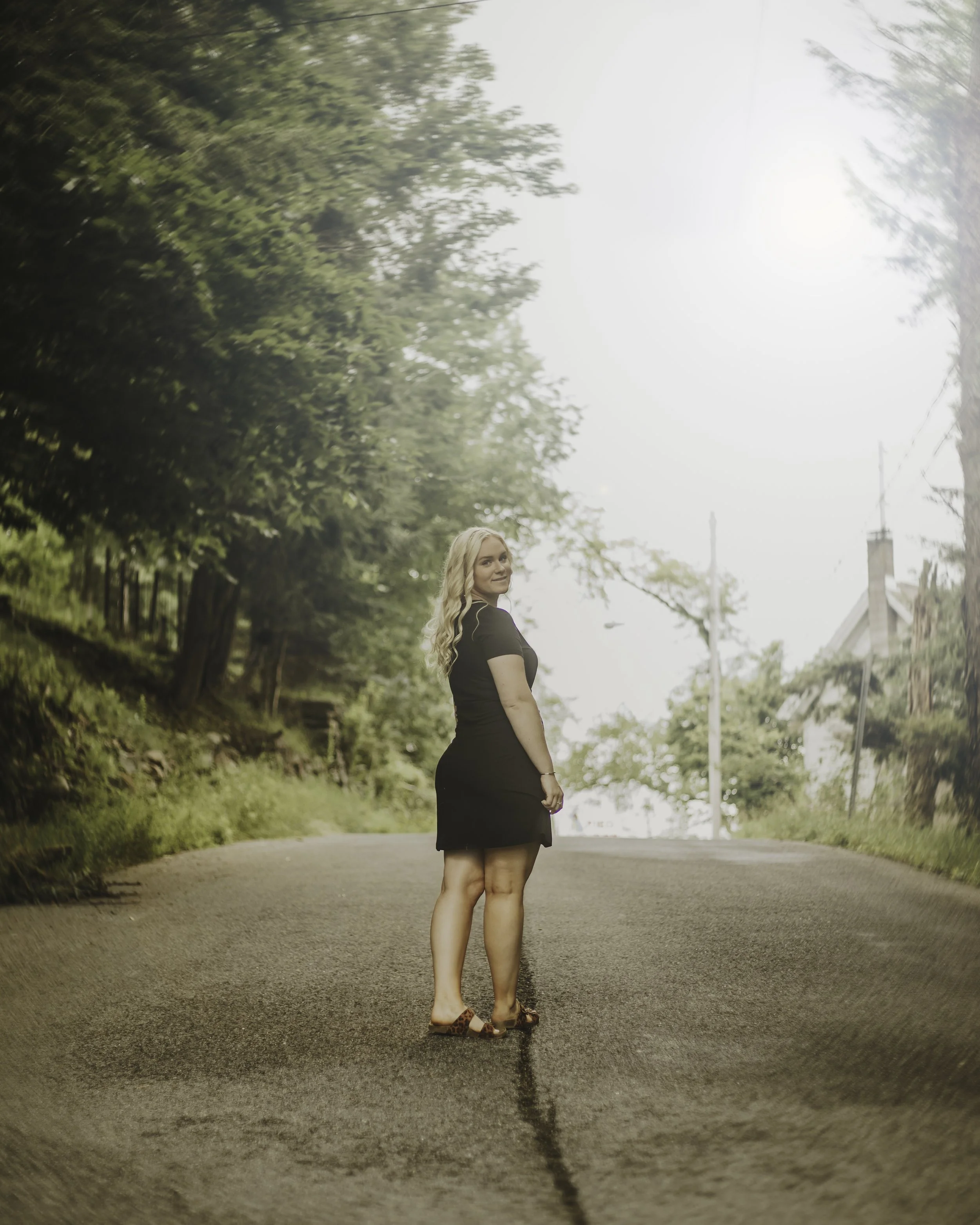 A woman in a black dress and sandals standing on an empty, paved street with greenery and trees on either side, under bright sunlight.