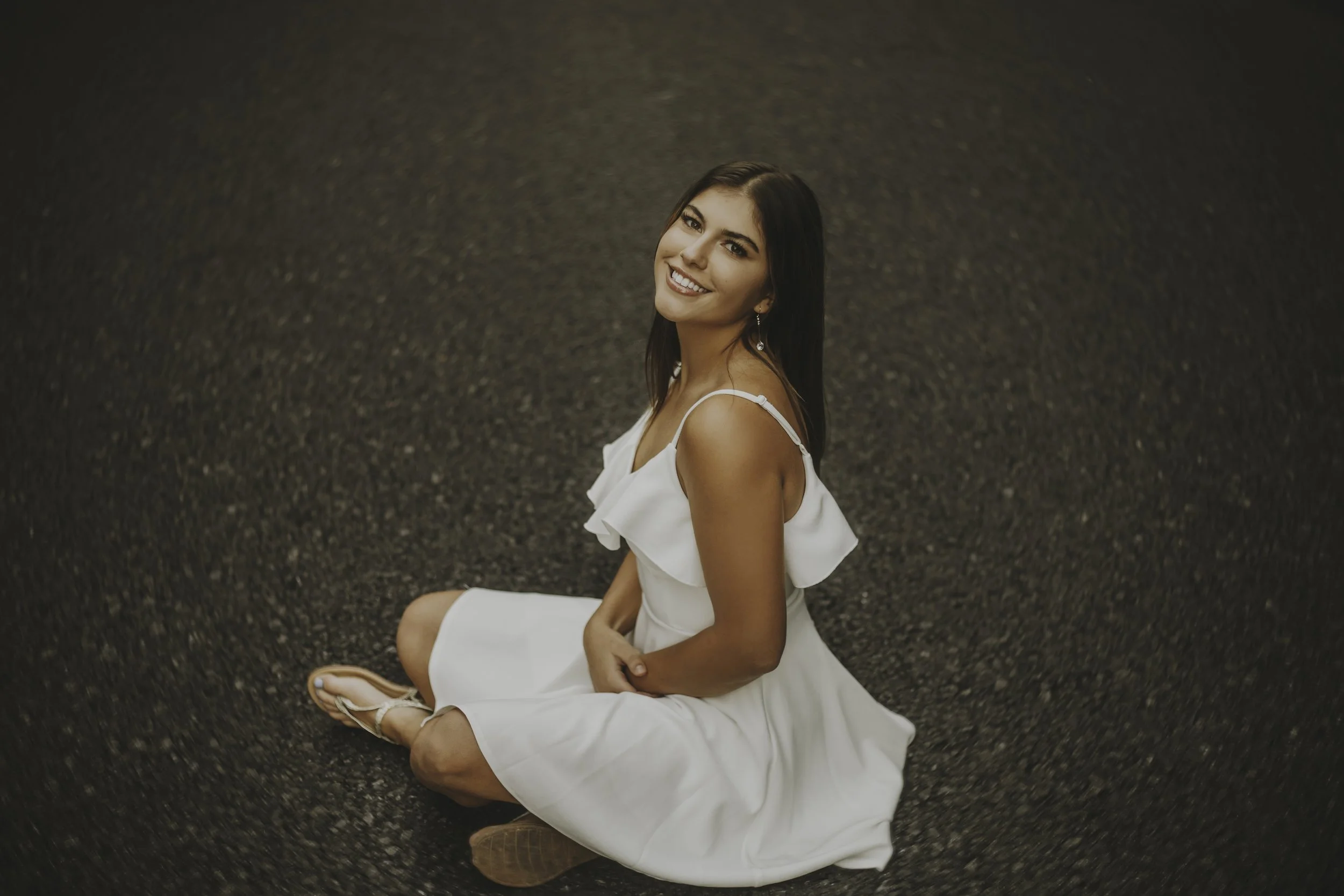 A woman in a white dress is sitting on a black asphalt surface, smiling at the camera.