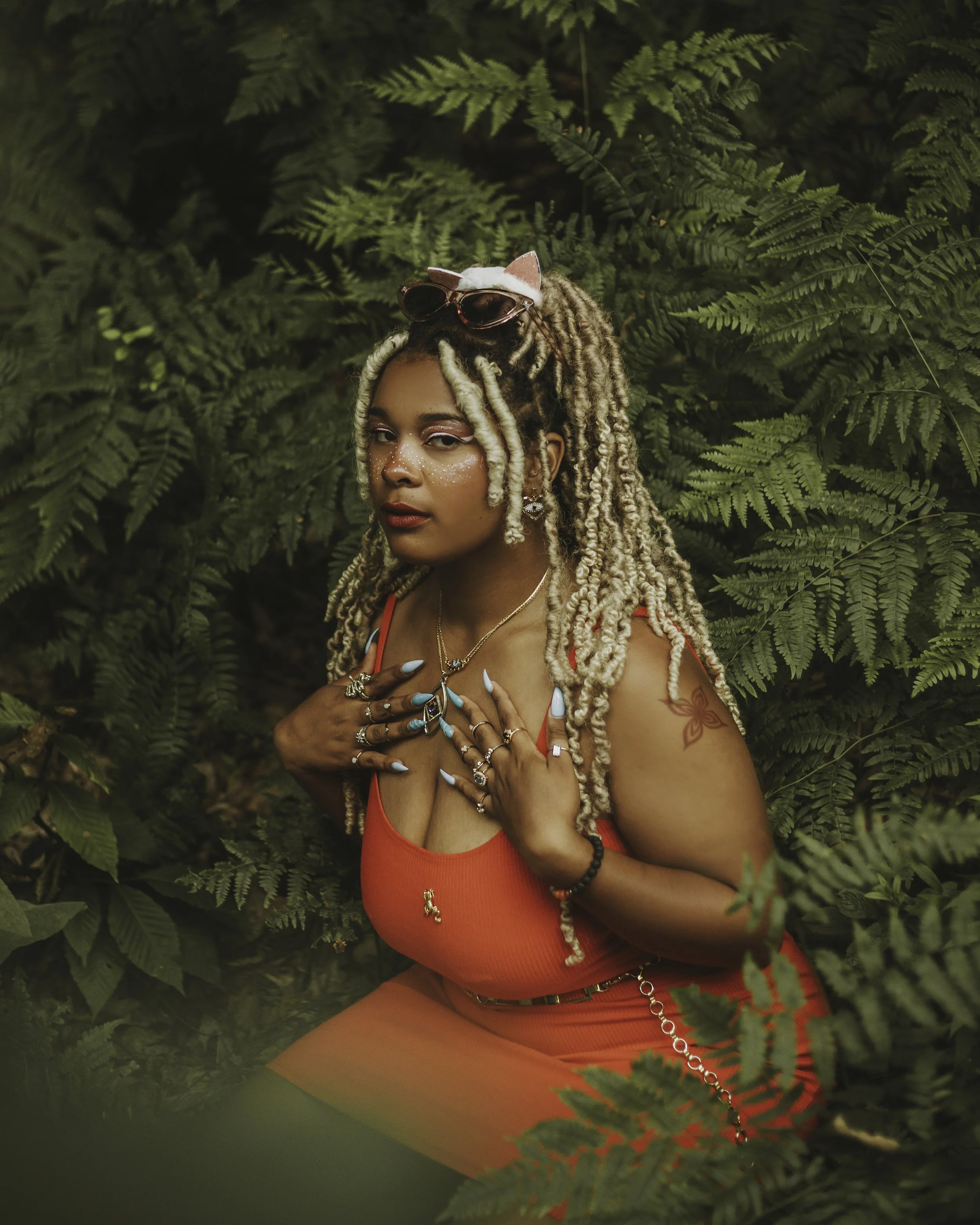 A woman with blonde dreadlocks, wearing red clothes, sunglasses on her head, and various jewelry, poses in front of green ferns.