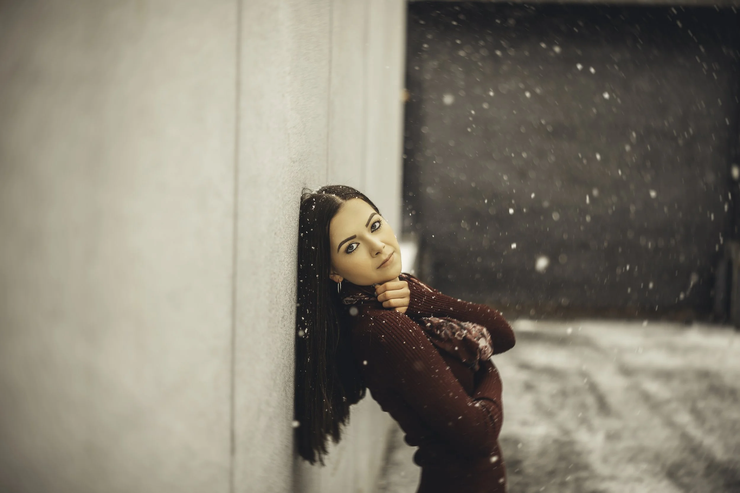 A woman with long dark hair and makeup, dressed in a red sweater and scarf, leaning against a wall in an outdoor snowing setting, looking towards the camera.