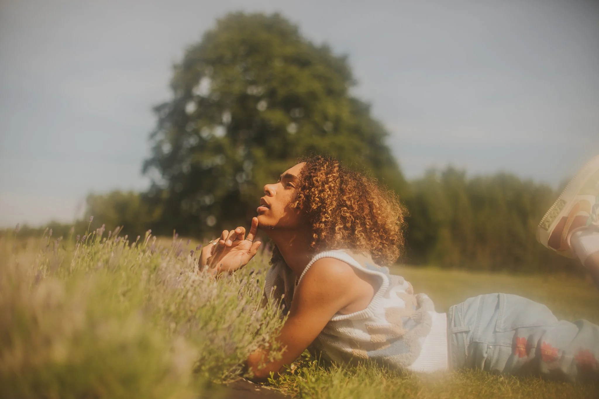 A woman with curly hair lying on her stomach in a grassy field, with a large tree in the background, looking thoughtfully upward.
