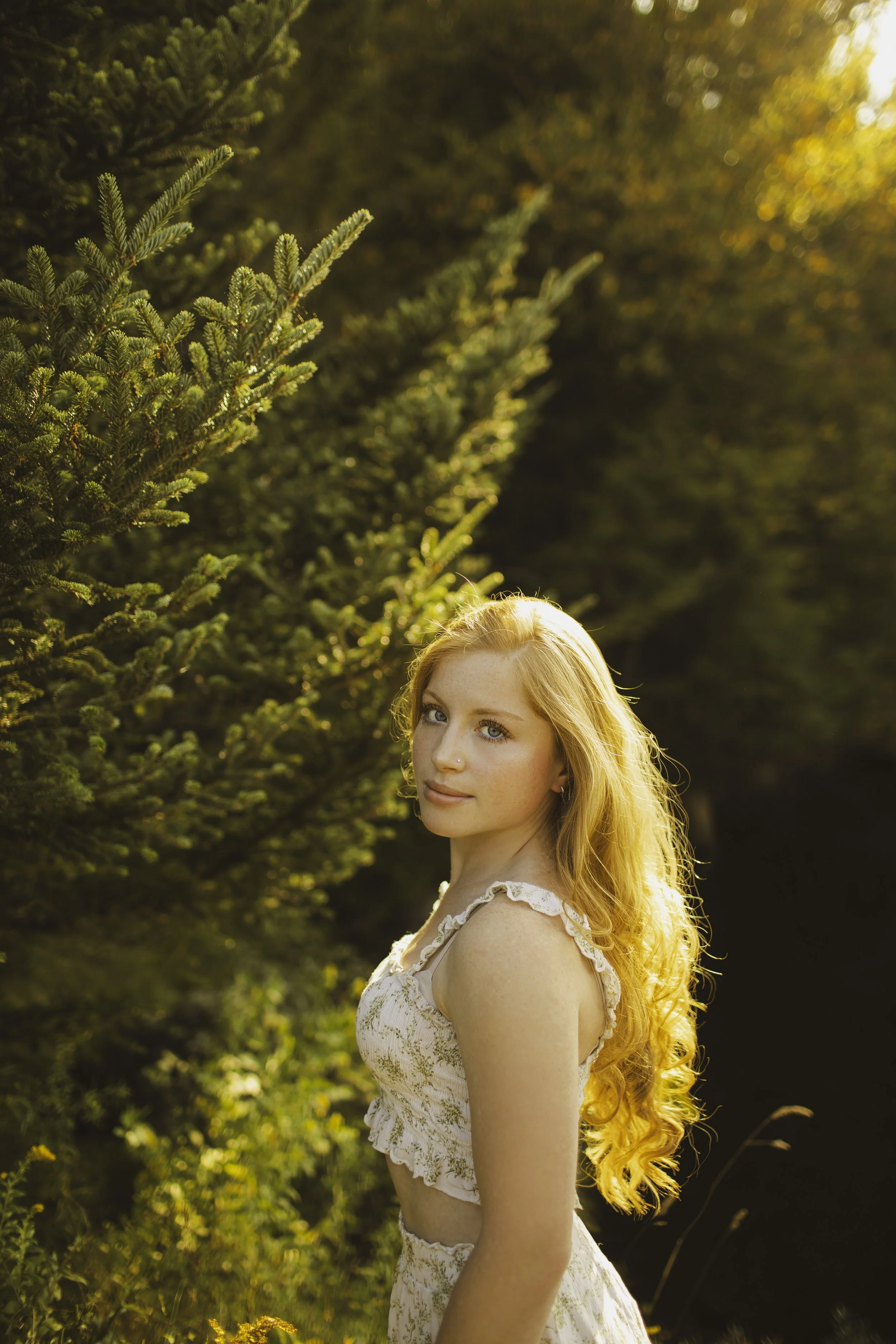 A young woman with long, curly red hair standing outdoors in a forest during sunset, wearing a sleeveless white dress with ruffles, looking at the camera.