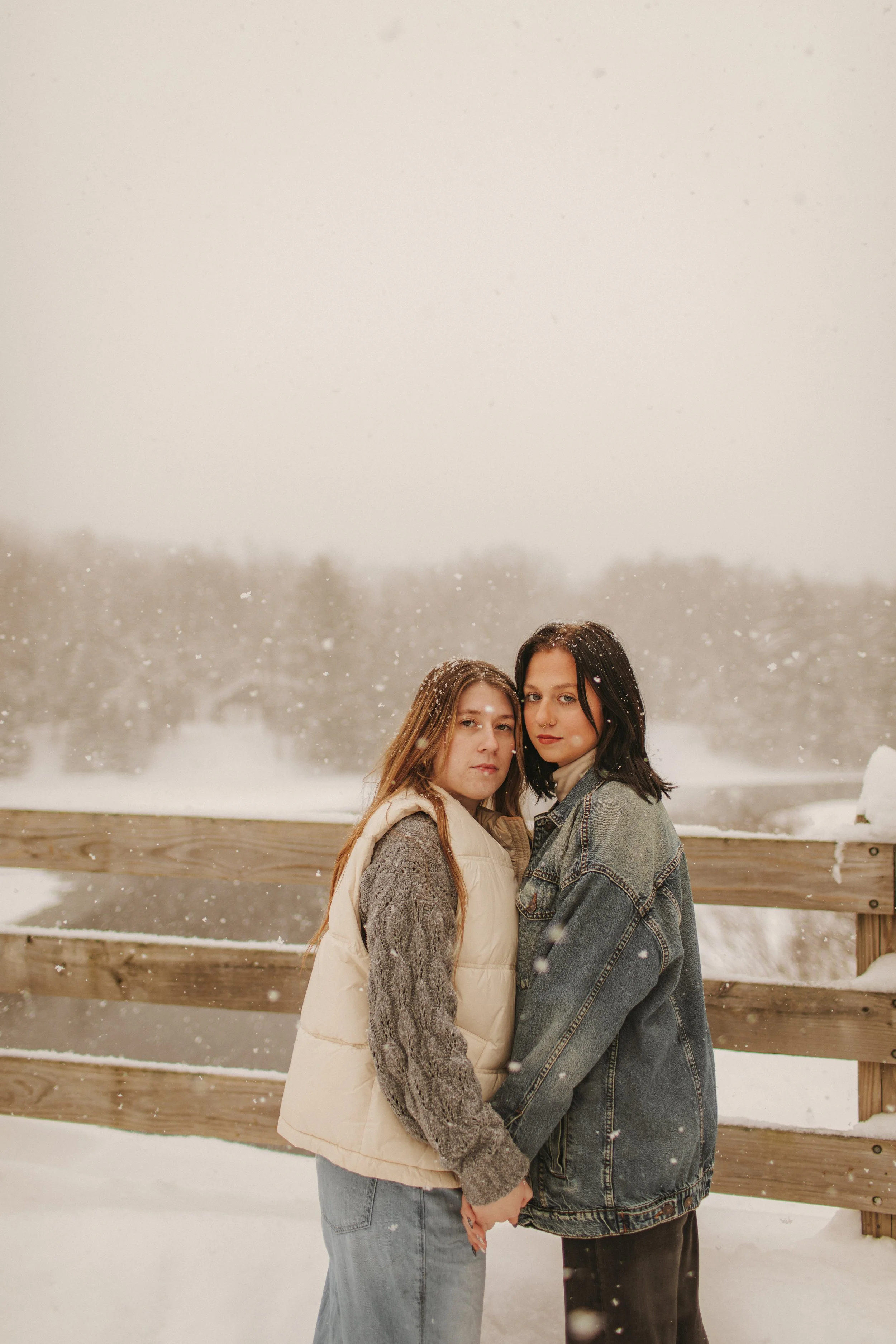 Two women stand close together on a snowy day near a wooden fence, holding hands and looking at the camera.