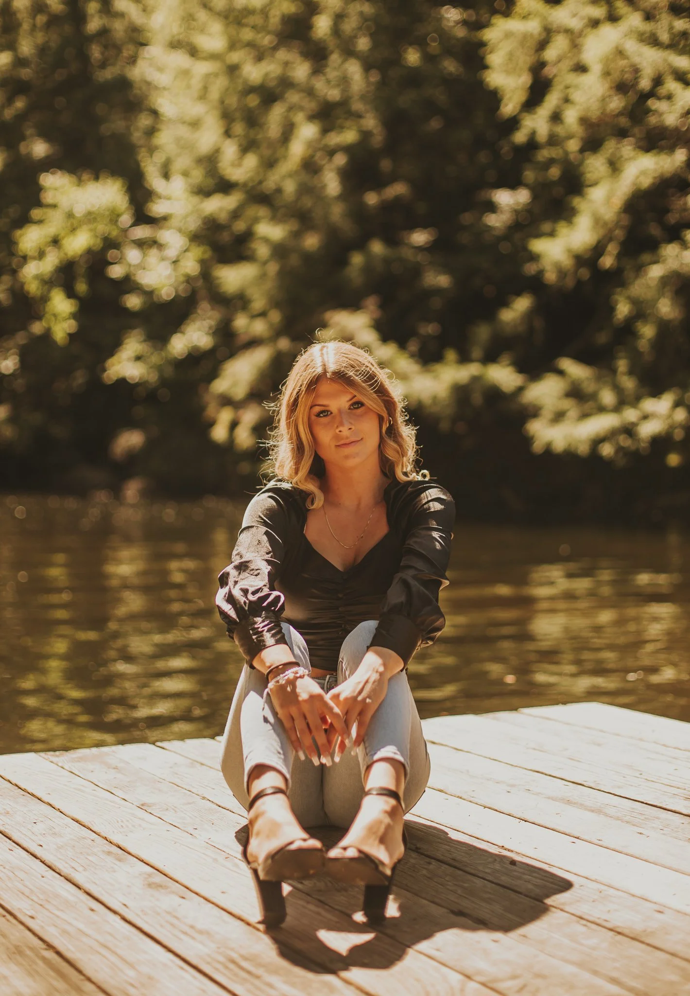 A young woman sitting on a wooden dock by the water, with trees in the background, wearing a black top, light-colored jeans, and sandals, looking at the camera.