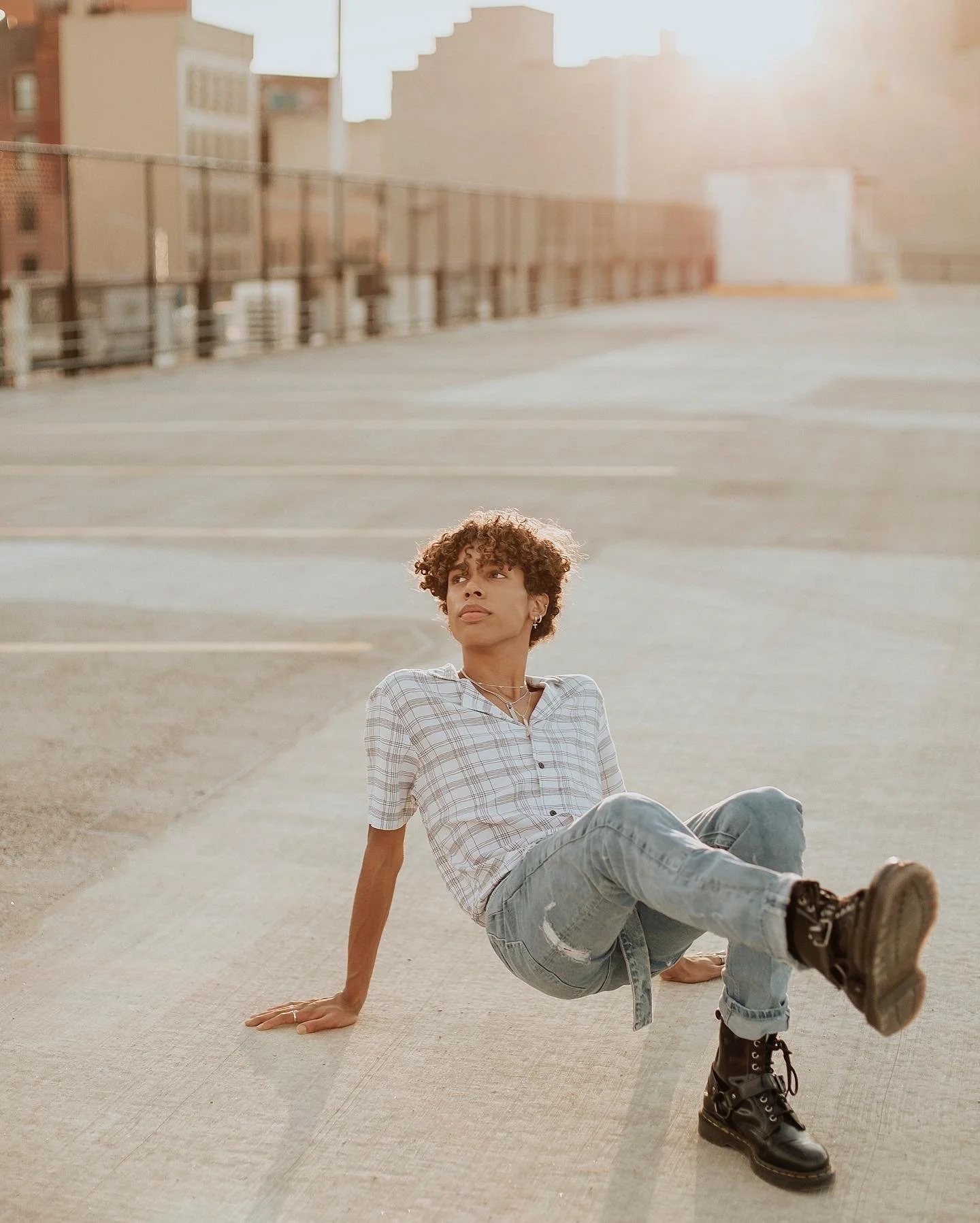 A woman with curly hair sitting on a parking garage rooftop during sunset, wearing a checkered shirt, ripped jeans, and black combat boots, with city buildings in the background.