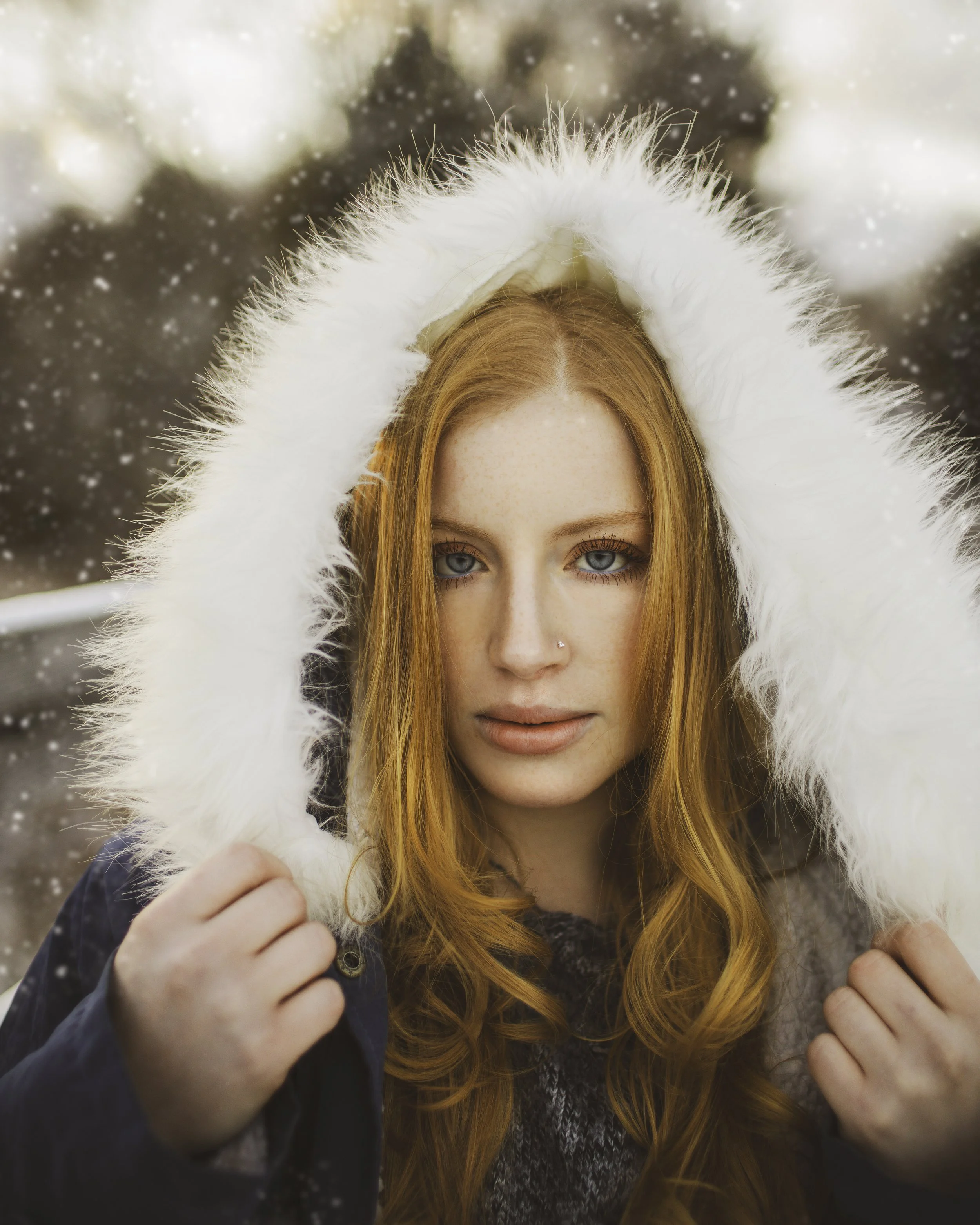 A red-haired woman in a winter coat with a white fur-lined hood, held with both hands, outdoors with falling snow and blurred trees in the background.