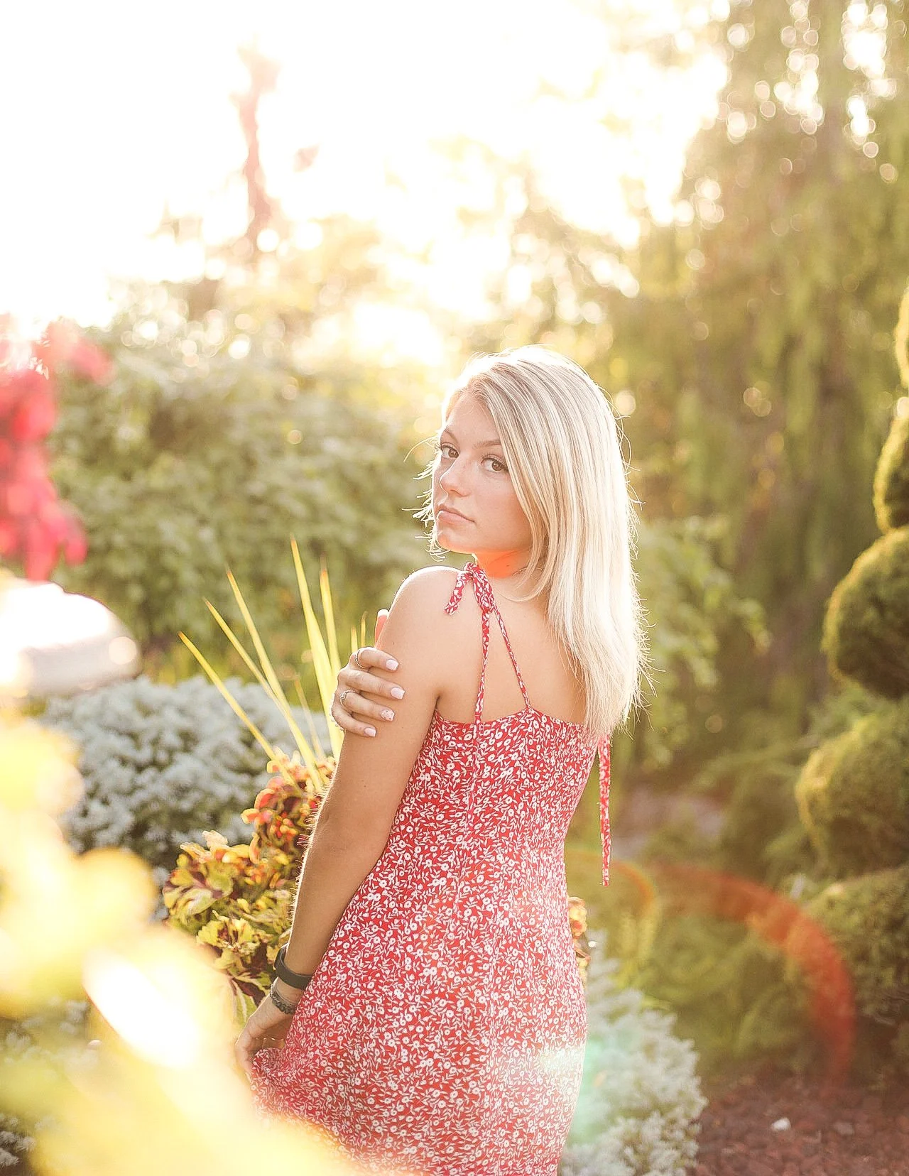 A young woman with blonde hair wearing a red floral dress standing in a sunlit garden surrounded by plants and flowers.