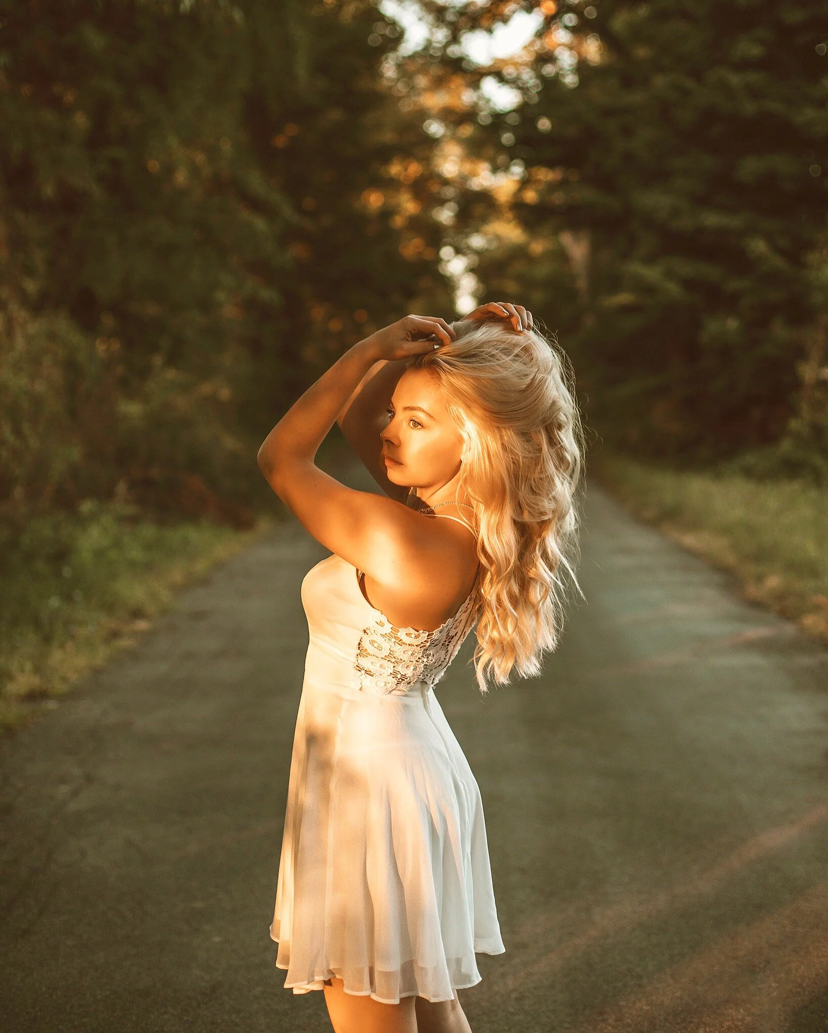 A woman with long blonde hair in a white dress, standing on a rural road at sunset, looking over her shoulder.
