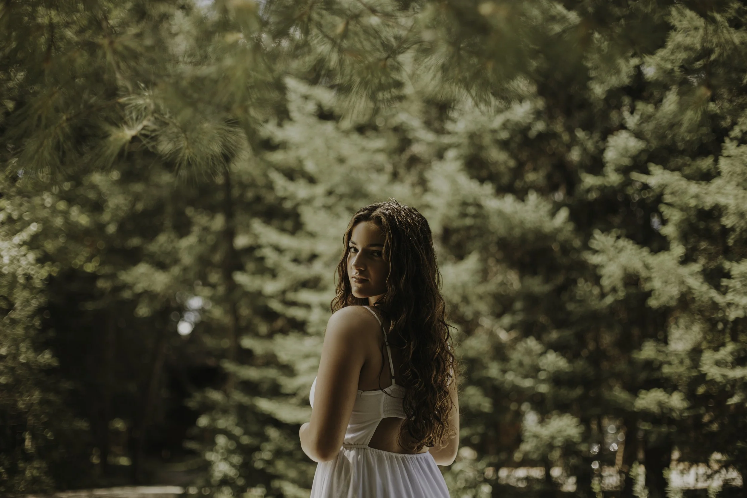 A young woman with long curly hair stands outdoors amidst trees, wearing a white dress and looking at the camera.