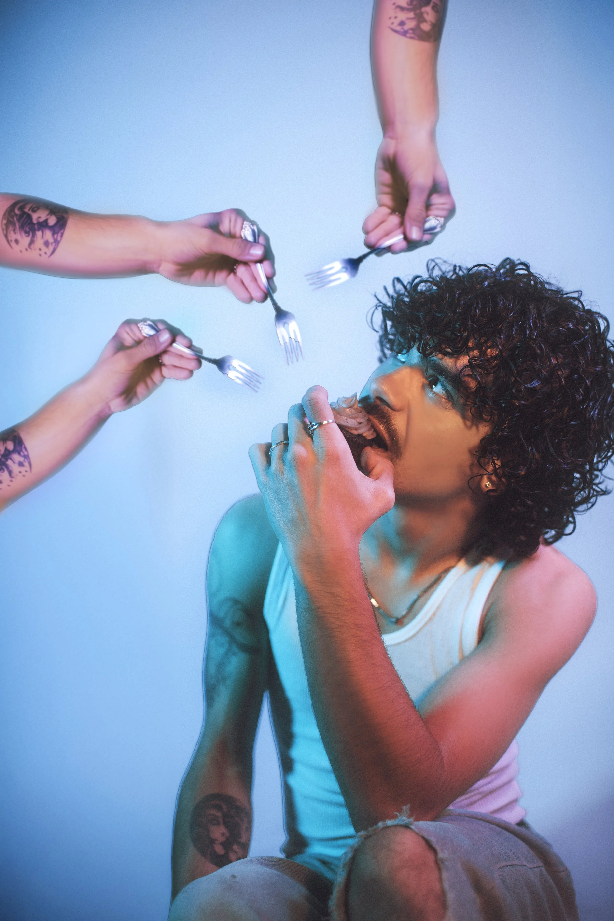 Person with curly hair wearing a white tank top, sitting and looking up as three hands with tattoos hold forks, feeding them food.