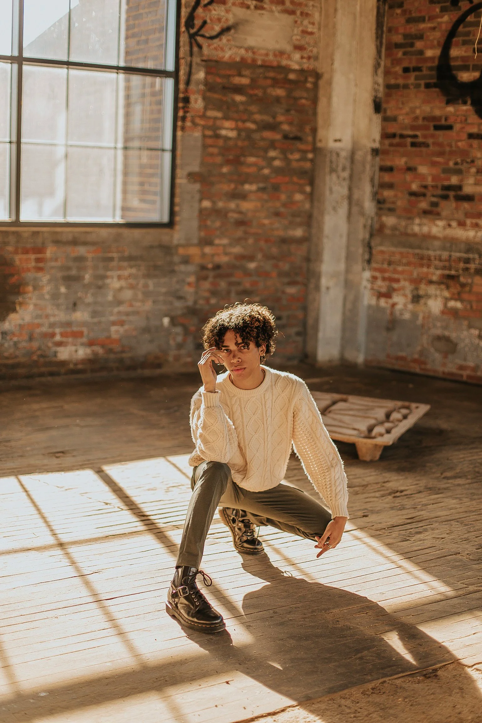 A young woman with curly hair squatting inside a rustic, industrial-style room with large windows, brick walls, and wooden floors. She is wearing a white knitted sweater, gray pants, and black leather boots, with sunlight casting shadows on the floor.