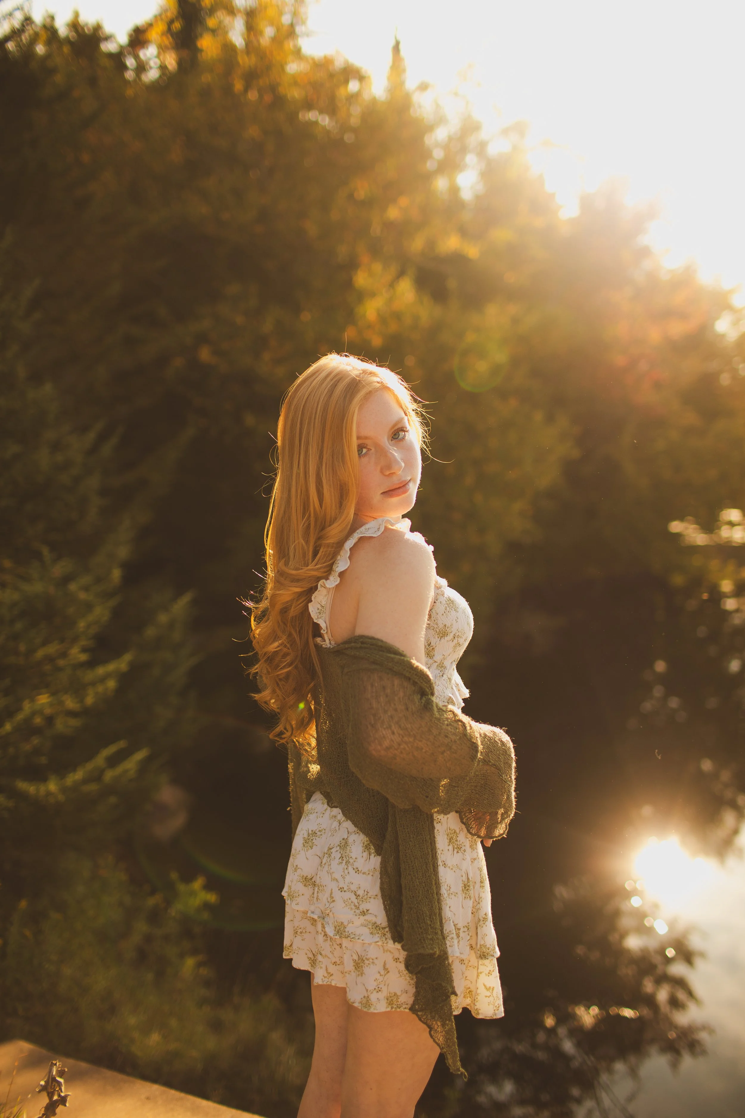 A young woman with long red hair standing outdoors in front of a body of water, illuminated by warm, golden sunset light, surrounded by trees with autumn leaves.