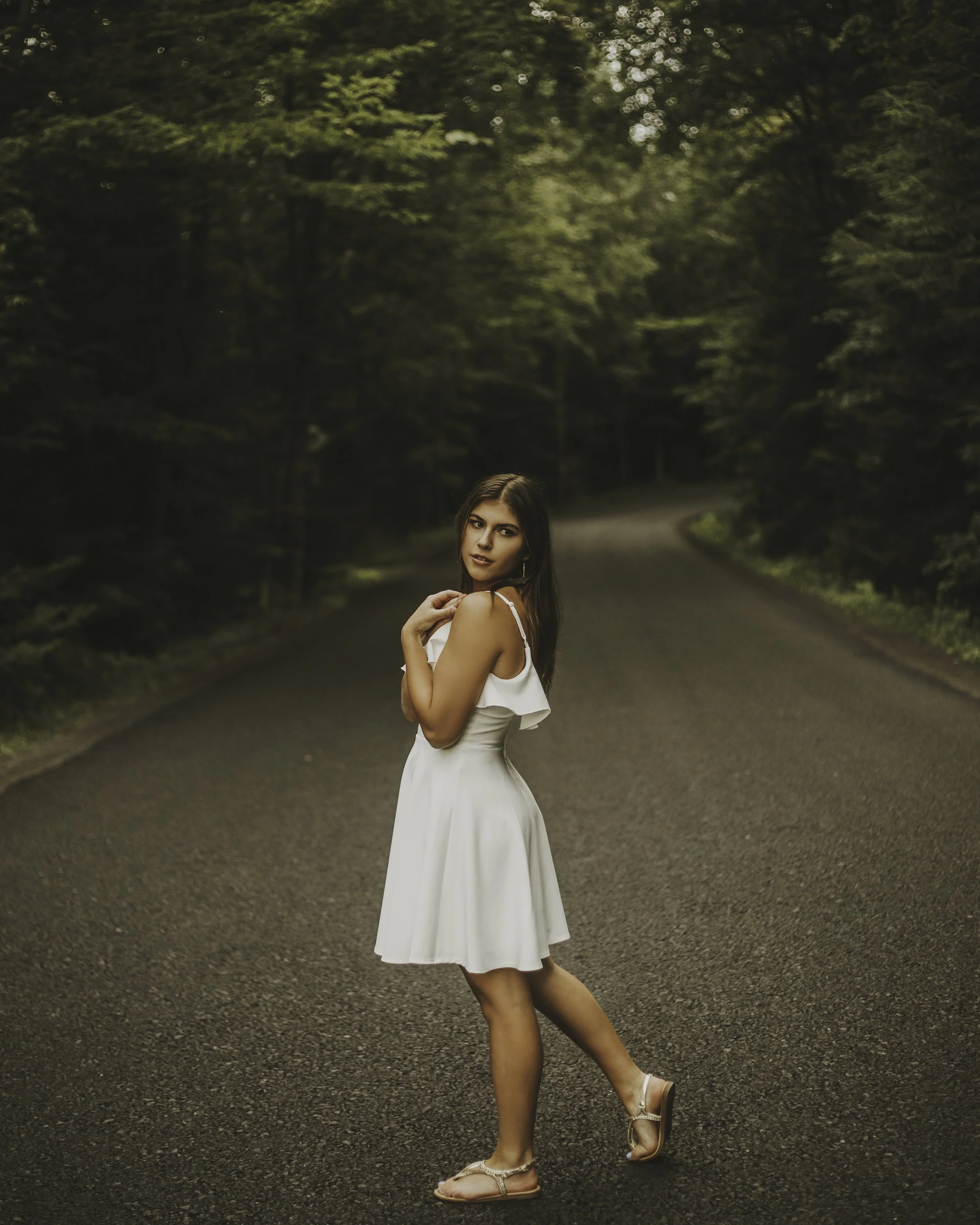 A young woman in a white dress and sandals standing on an empty road surrounded by lush green trees.