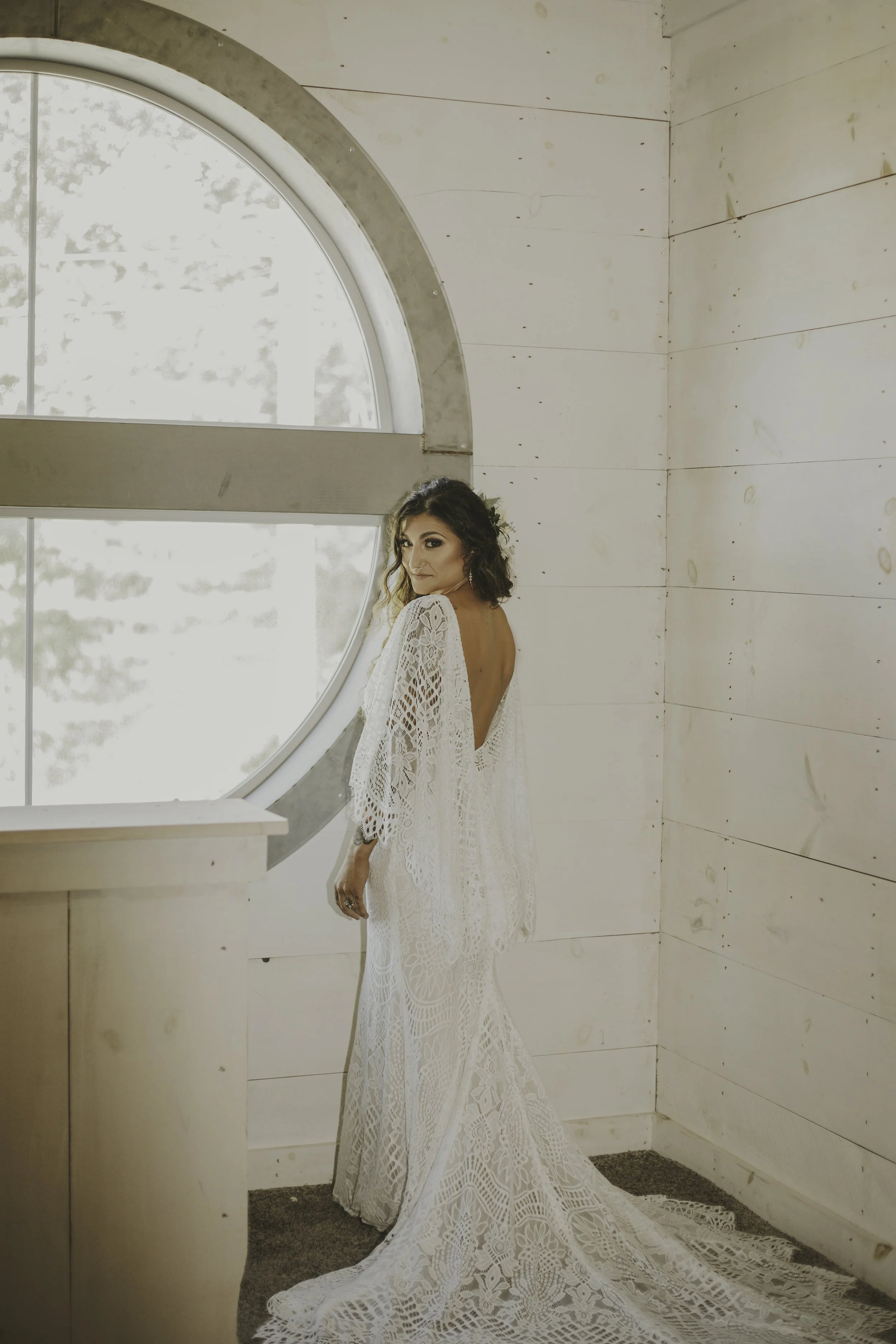A woman in an intricate white lace wedding dress, standing indoors near a large round window with a wooden wall background.