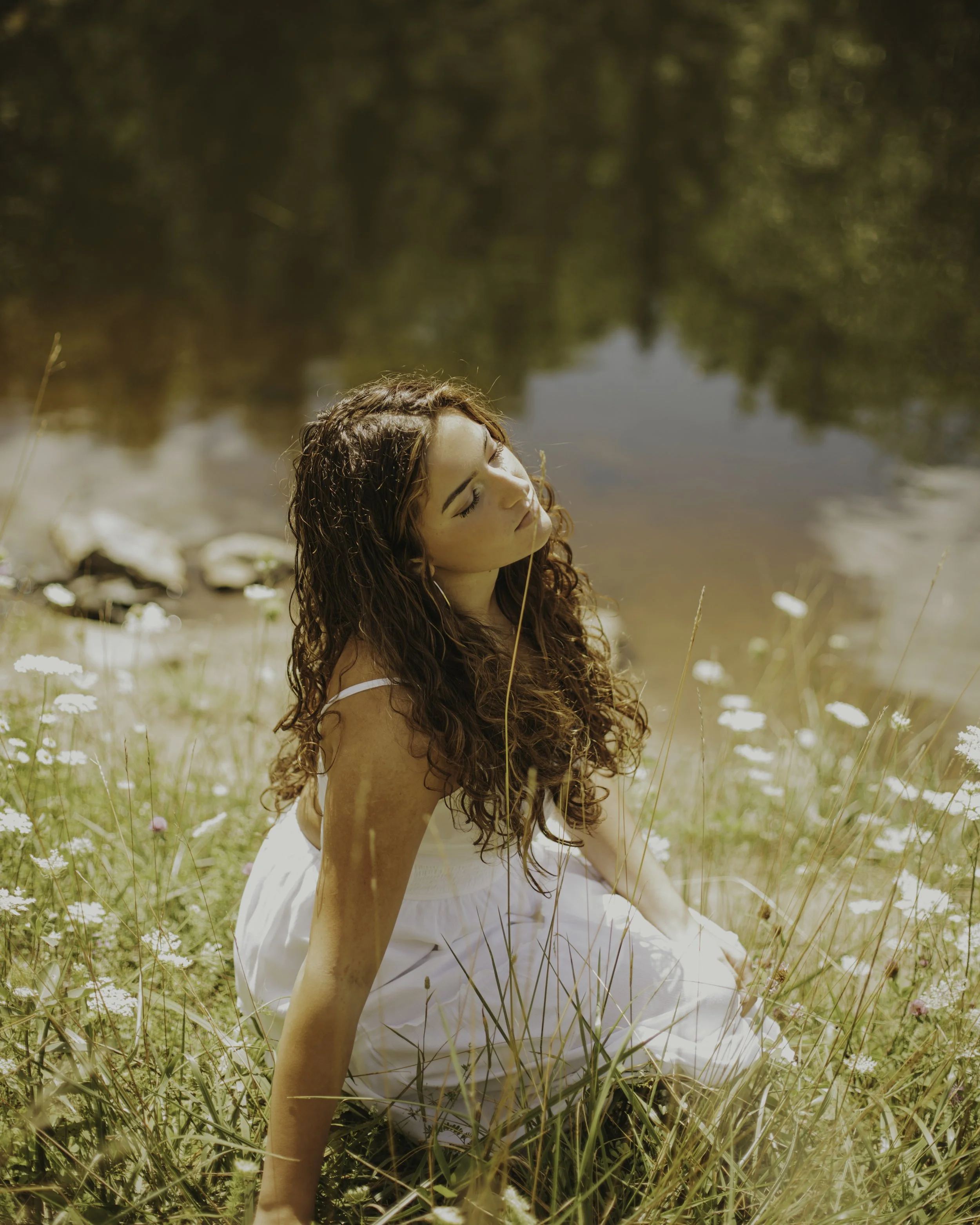A woman with long, curly hair sitting in a field of tall grasses and wildflowers near a lake on a sunny day.