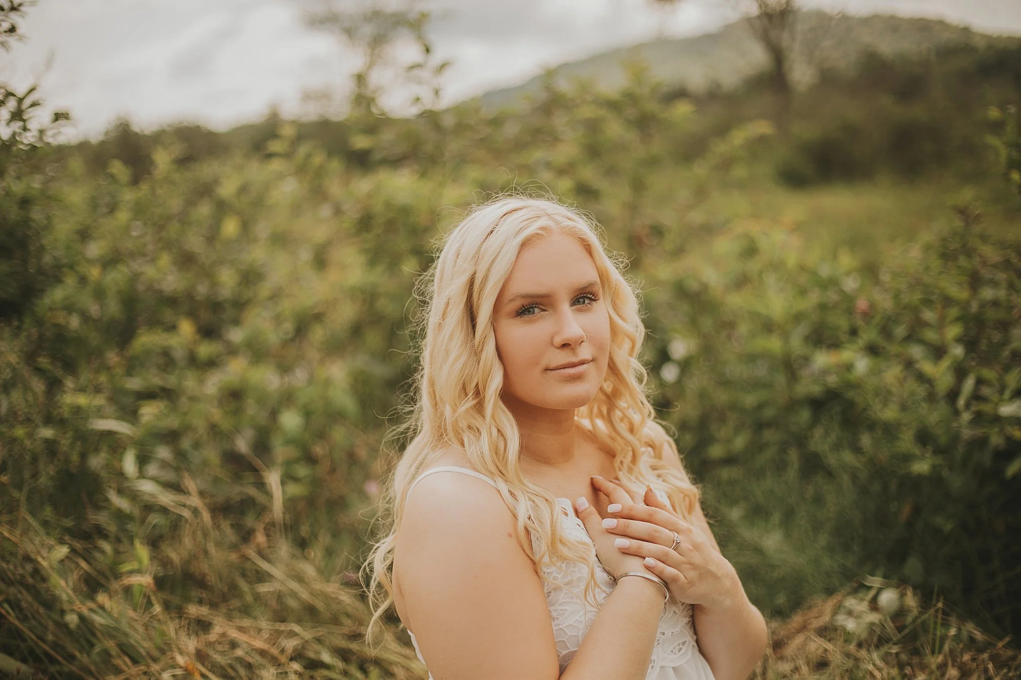 A young woman with long blonde curly hair wearing a white dress standing outdoors in a natural setting with greenery and hills in the background, with her hands on her chest.