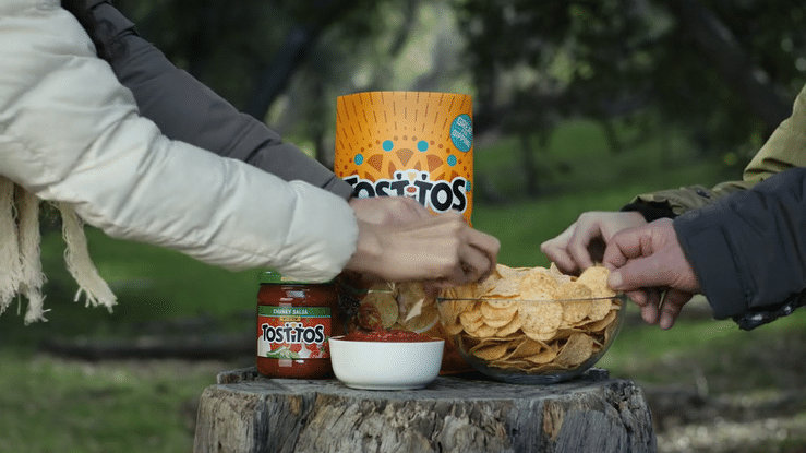 Two people reaching for chips from a bowl on a tree stump, with Tostitos chips, salsa, and dip on the stump, outdoors in a park.