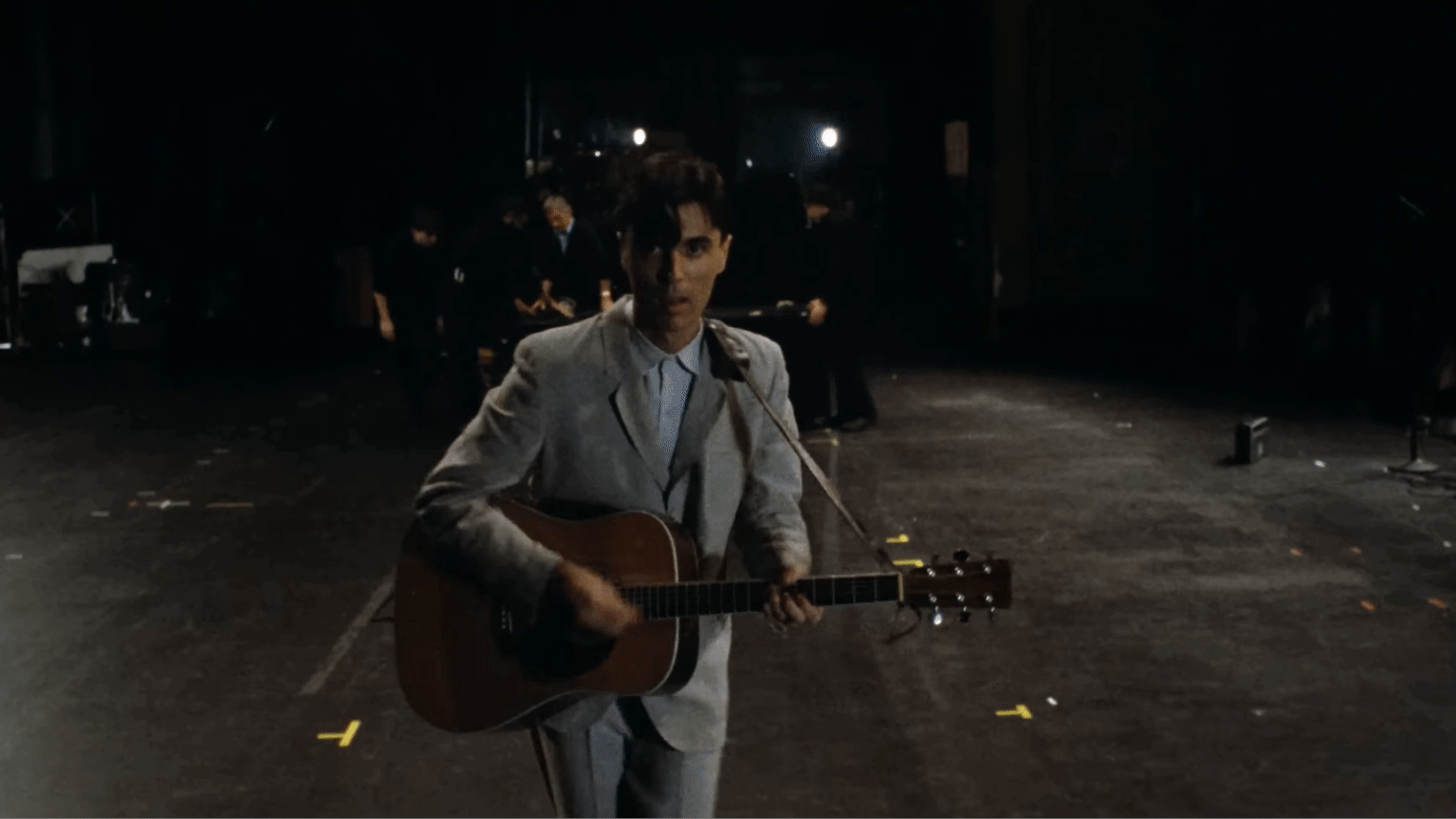 A man in a light-colored suit playing an acoustic guitar in a dimly lit room.