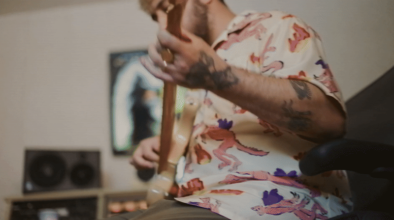 A person with tattoos on their arm, wearing a floral shirt, sitting in front of a TV and playing an electric guitar.