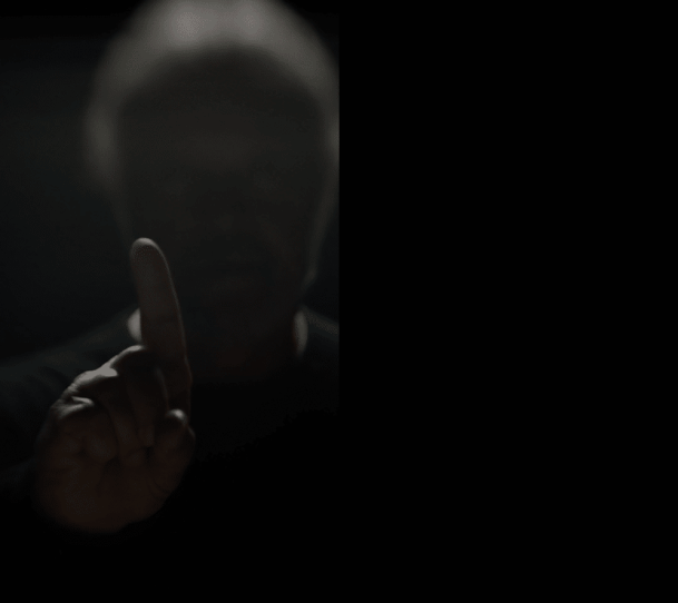 Person with gray hair raising their index finger in front of their face, image taken in low light with partial shadow.