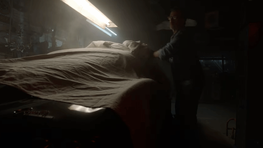 Person making a bed in a dimly lit room with a fluorescent light, possibly a workshop or garage, surrounded by various tools and equipment.