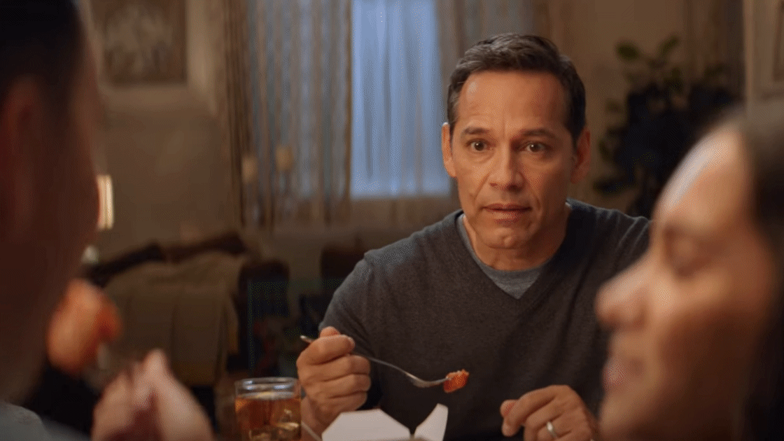 A middle-aged man with short dark hair eating a meal at a table with two others in a cozy, warmly lit room.