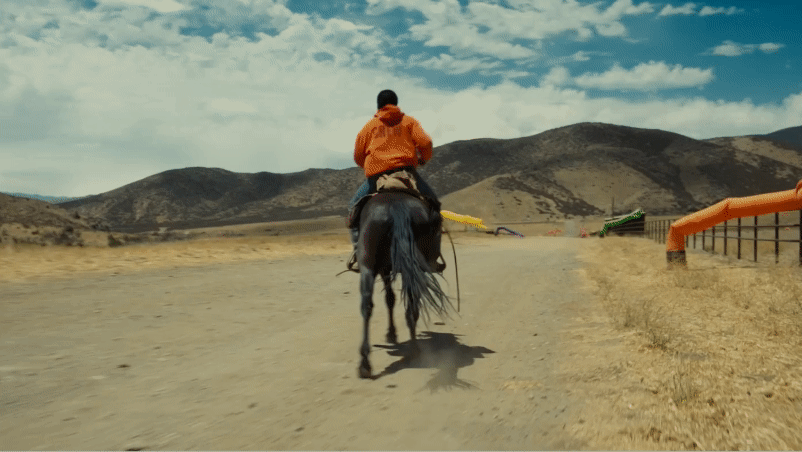 A person riding a horse on a dirt trail through a mountainous area with blue sky and scattered clouds.