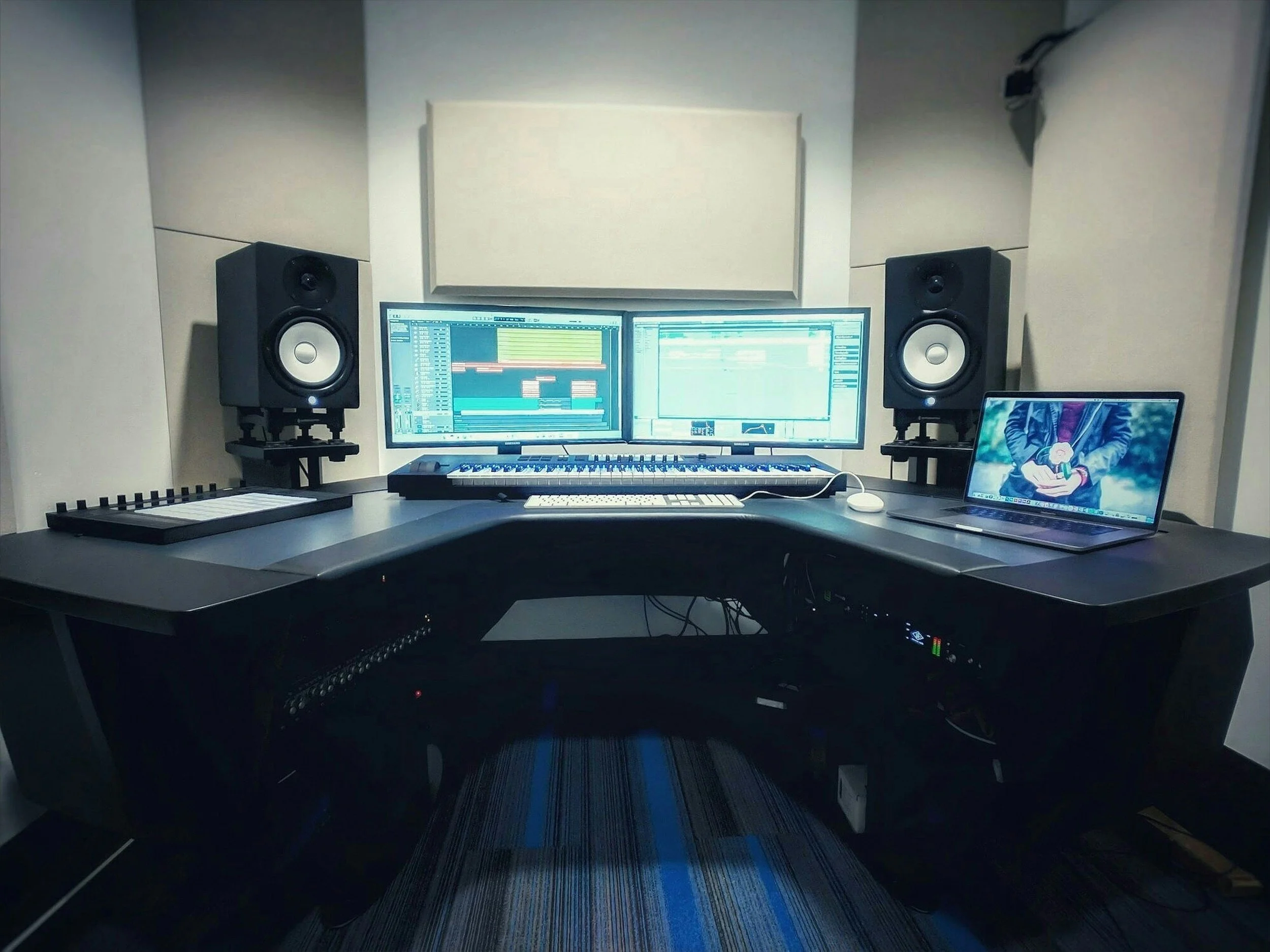 Music production studio setup with dual monitors displaying digital audio workstations, two black studio speakers on stands, a keyboard, a laptop showing a person holding a dog, and a MIDI controller on a black desk.